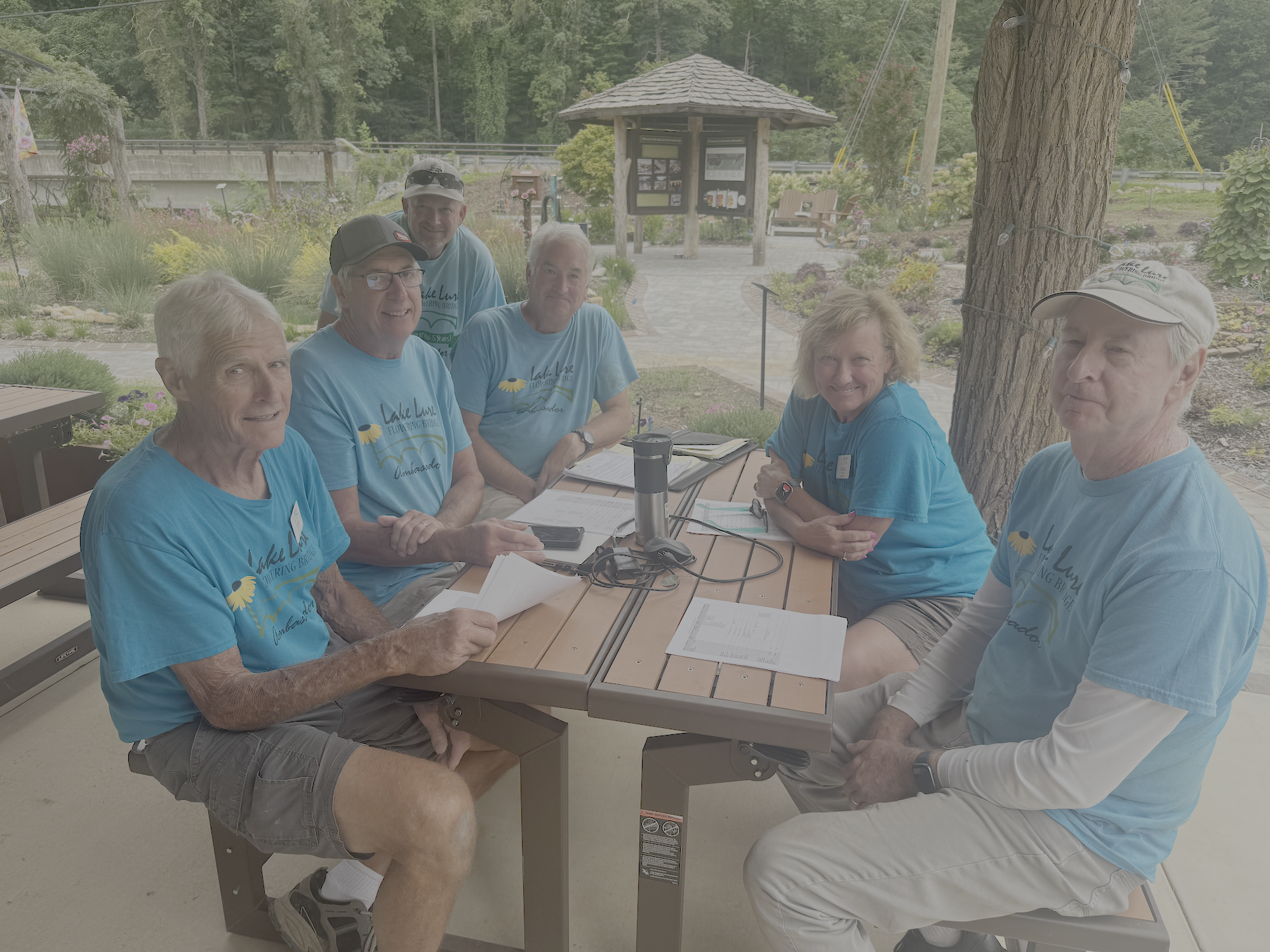 Six people sitting at a table outdoors, wearing matching blue t-shirts, smiling, with trees and a gazebo in the background.