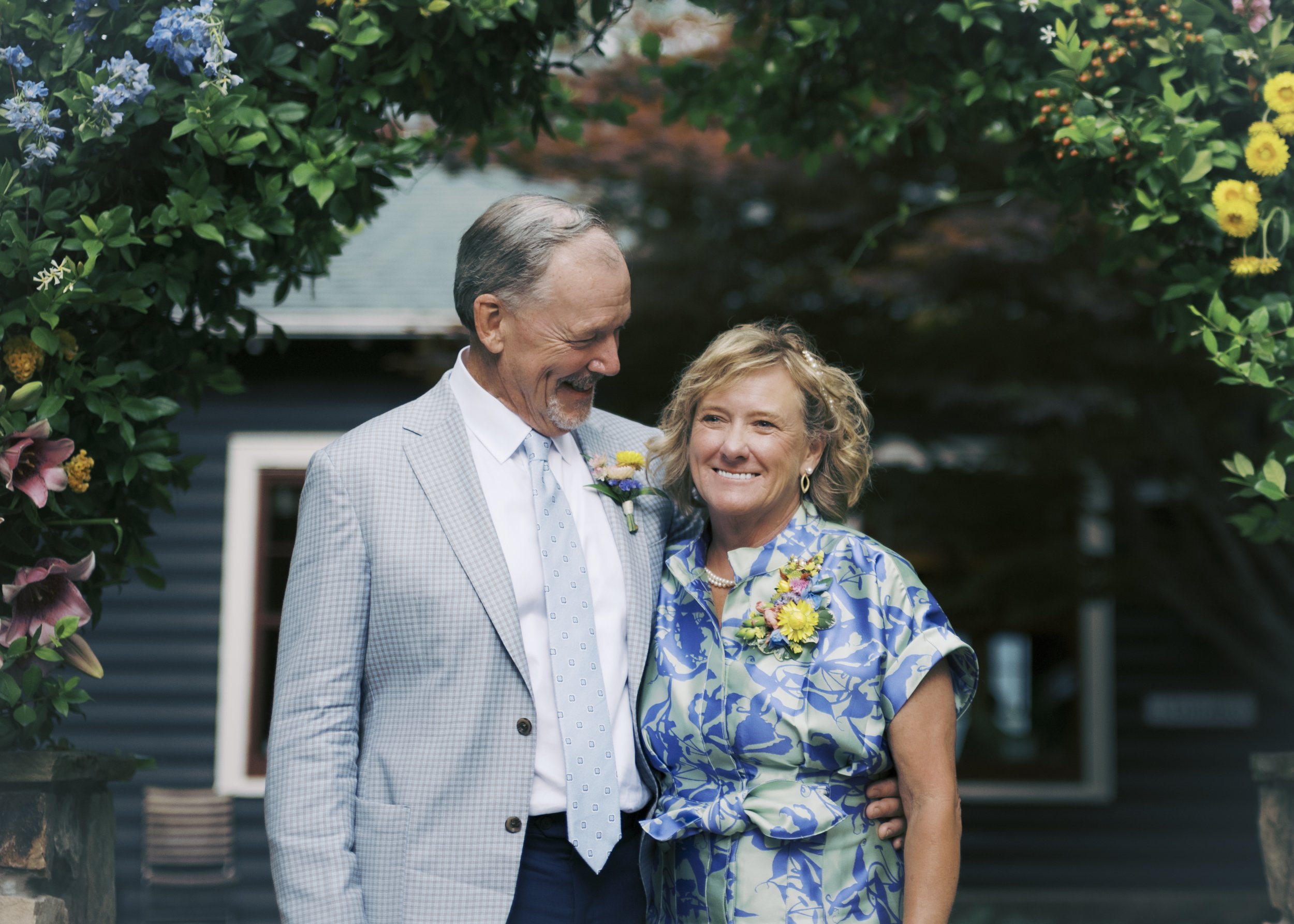 A happy elderly couple standing outdoors among lush greenery and flowers, smiling and embracing each other during a sunny day.
