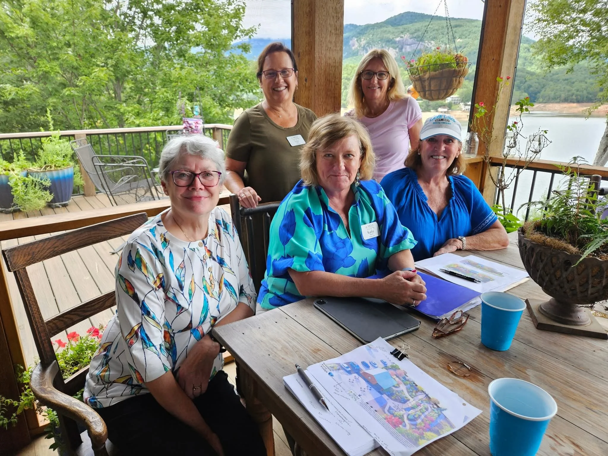 A group of six women gathered around a wooden table on a covered porch, with greenery and mountain views in the background. They are smiling and appear to be at a meeting or social gathering, with papers, notebooks, and a map on the table.