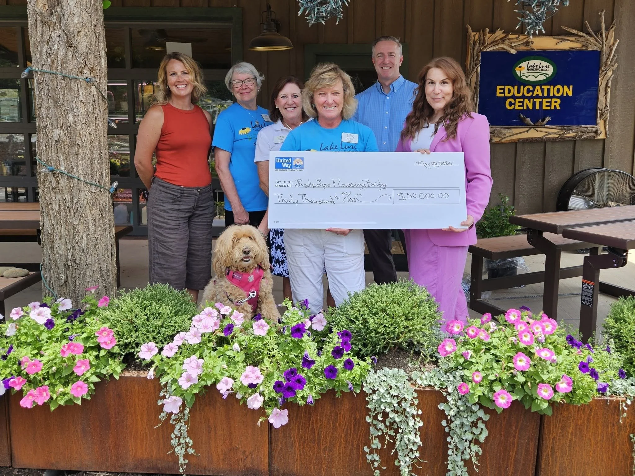 Group of six people standing outdoors in front of a wooden building and sign that reads 'Education Center.' They are holding a large check for $30,000 made out to Lake Luna Flowering Bridge. A dog sits in front of them, and there are colorful flowers in the foreground.