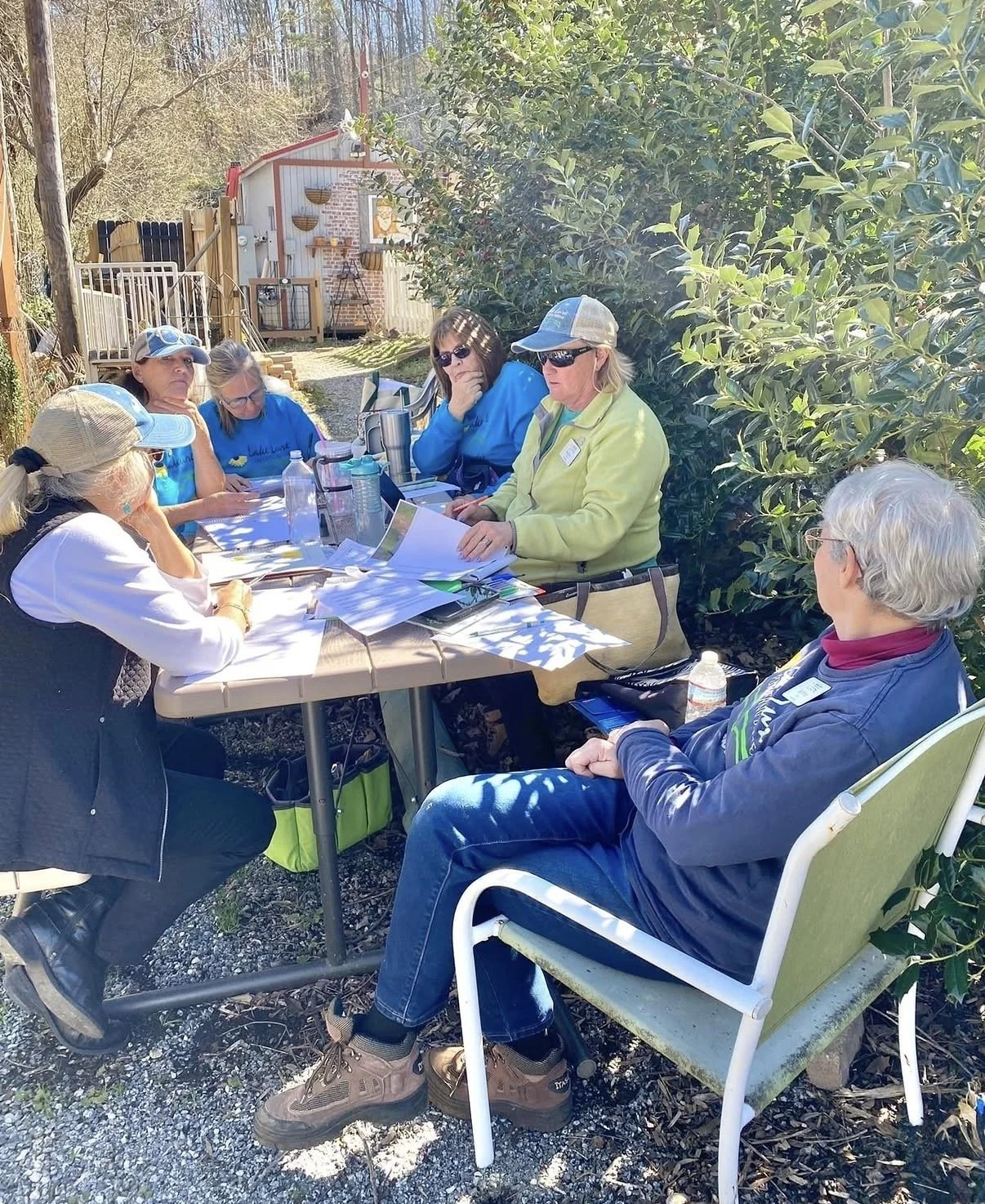Group of women sitting around a table outdoors, reading and discussing papers, surrounded by trees and sunlight.