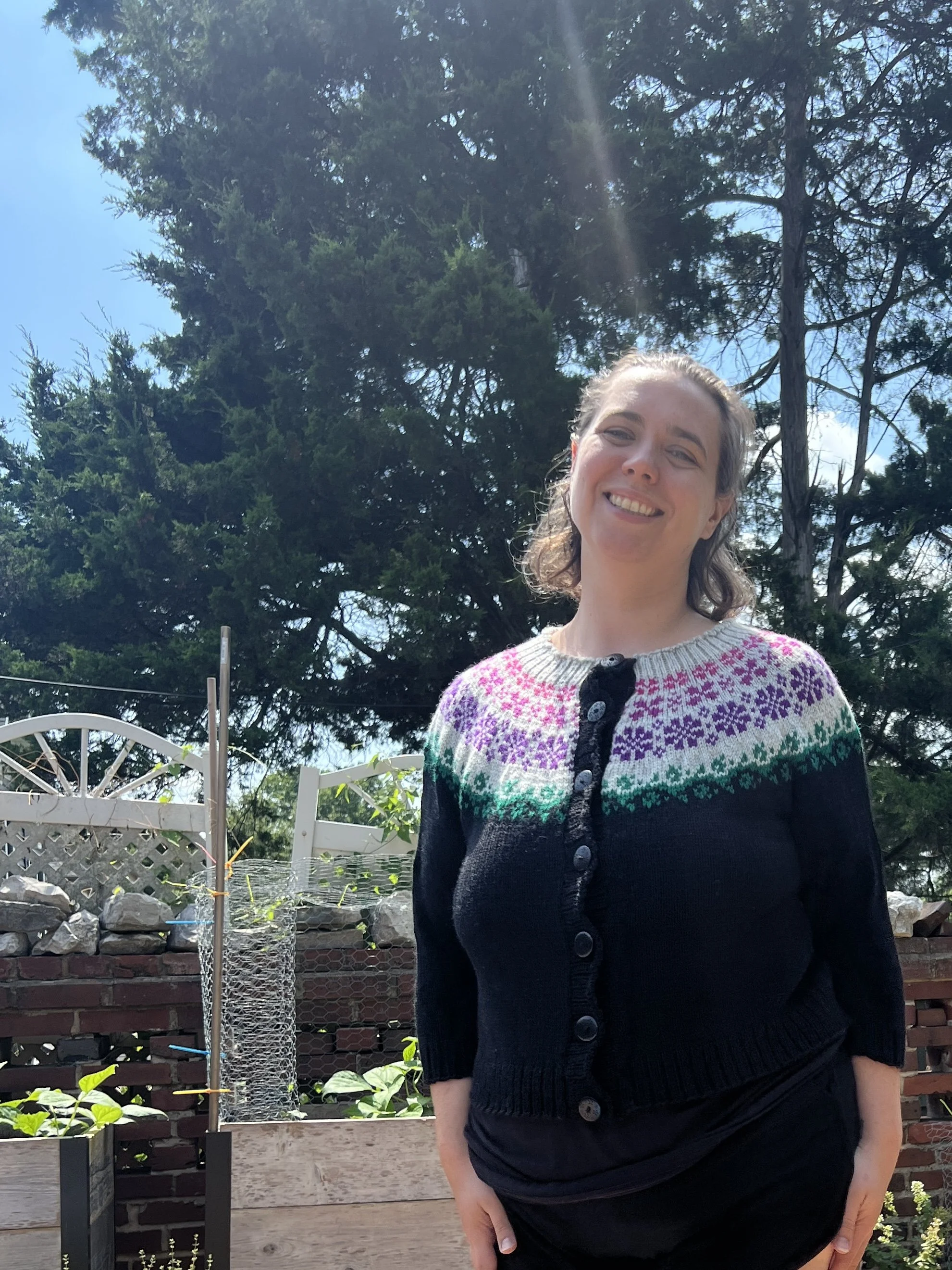 Katherine Smith, founder of Homespun Knits, wearing a colorful hand made knitted sweater, standing outdoors in a garden area with trees, plants, and a brick wall in the background.