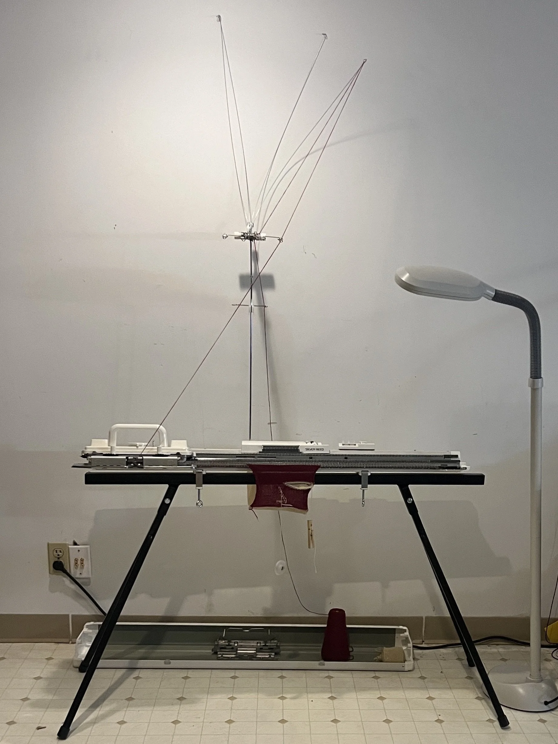My studio setup at Homespun Knits.  This is my standard gauge Silver Reed knitting machine mounted on a stand with yarn threaded through its needles, next to a floor lamp.