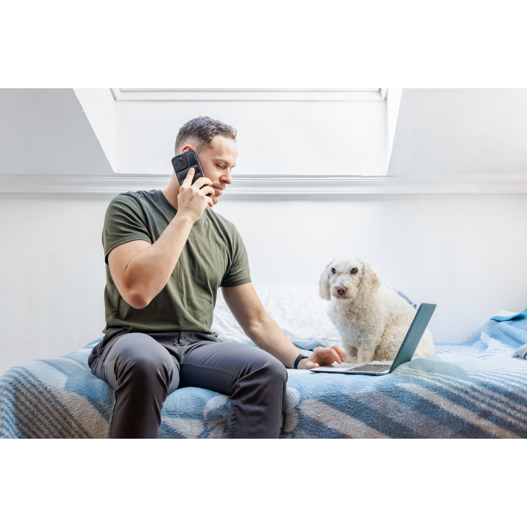 A man sitting on a bed, talking on his cellphone, with a laptop in front of him and a white poodle dog beside him. The room has a bright skylight window.