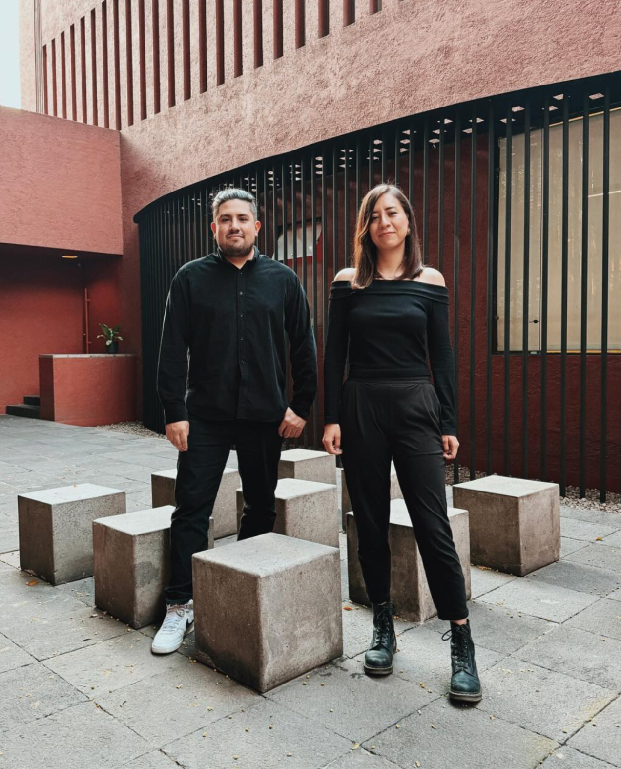 A man and woman standing outdoors in front of a modern red building with black metal fencing. The man is wearing a black shirt and pants, and the woman is in a black off-shoulder top and black pants. They are standing on a paved area surrounded by concrete blocks.