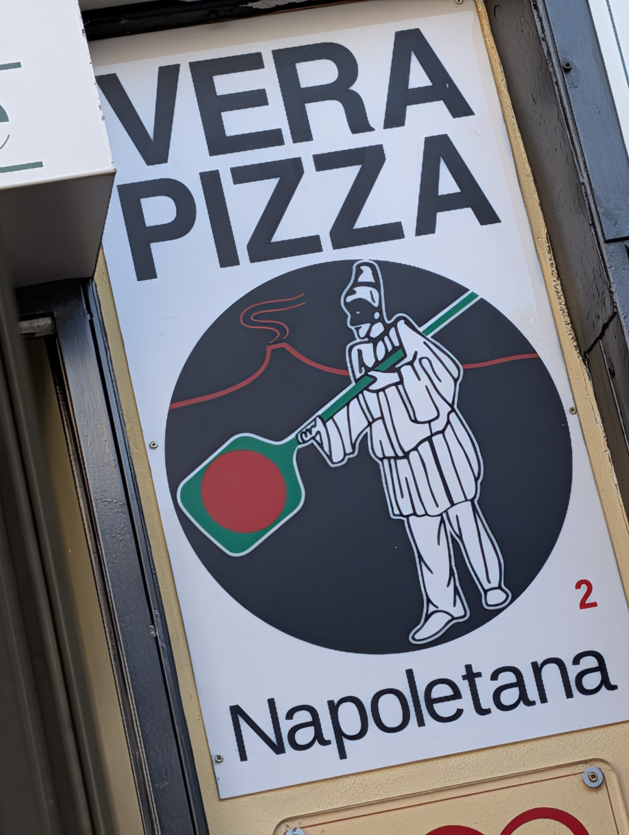 Schild mit dem Text "Vera Pizza", einem grafischen Symbol eines Kochs mit Schneebesen in der Hand, der eine Pizza hält, und einem rot-grün-weißen italienischen Flaggelement, das die italienische Pizzatradition repräsentiert, an einem Restaurant oder Pizzastand in Neapel.