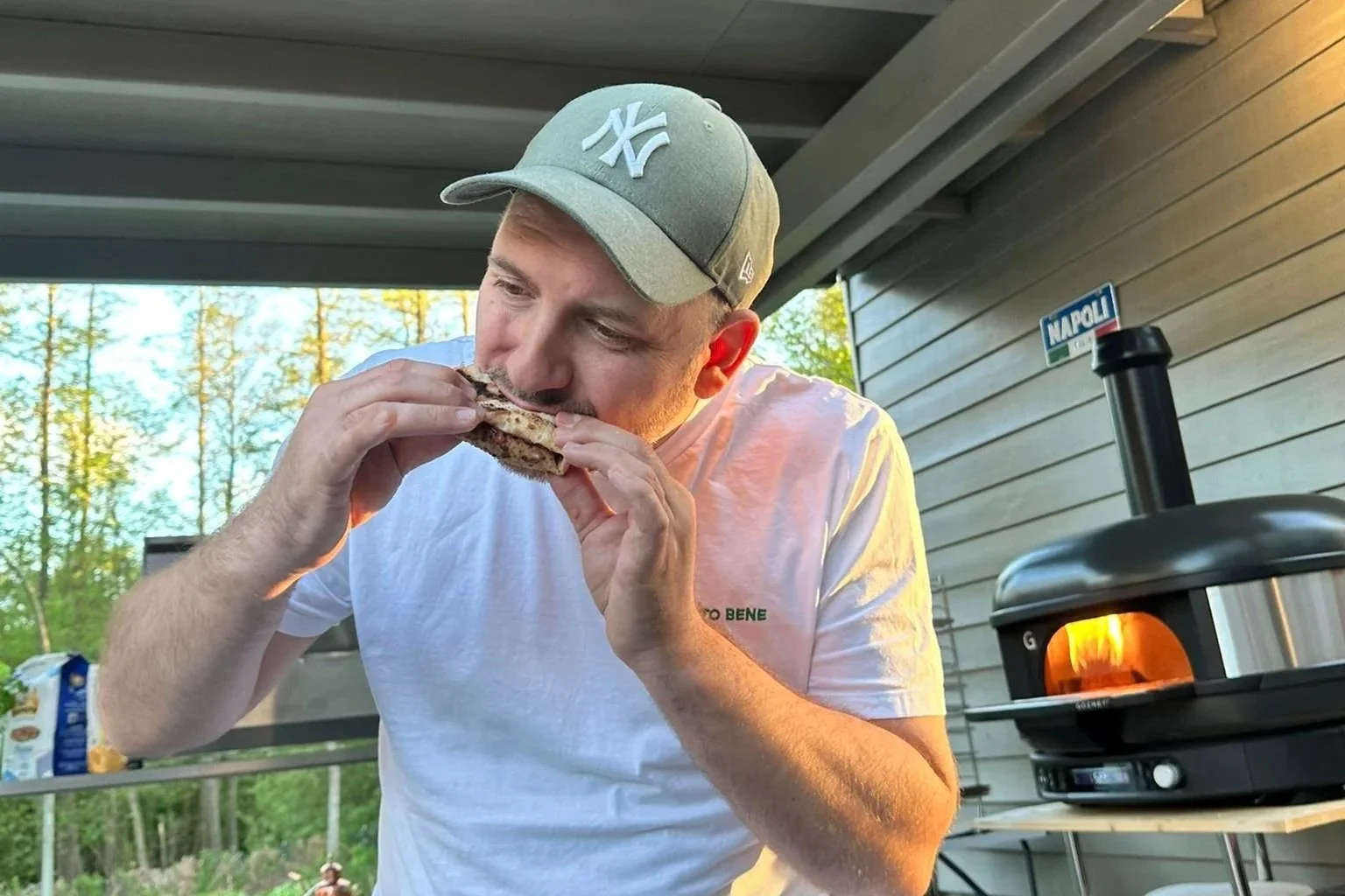 Mann beim Essen eines Sandwiches im Freien, neben einem Pizzaofen, mit Baum im Hintergrund.
