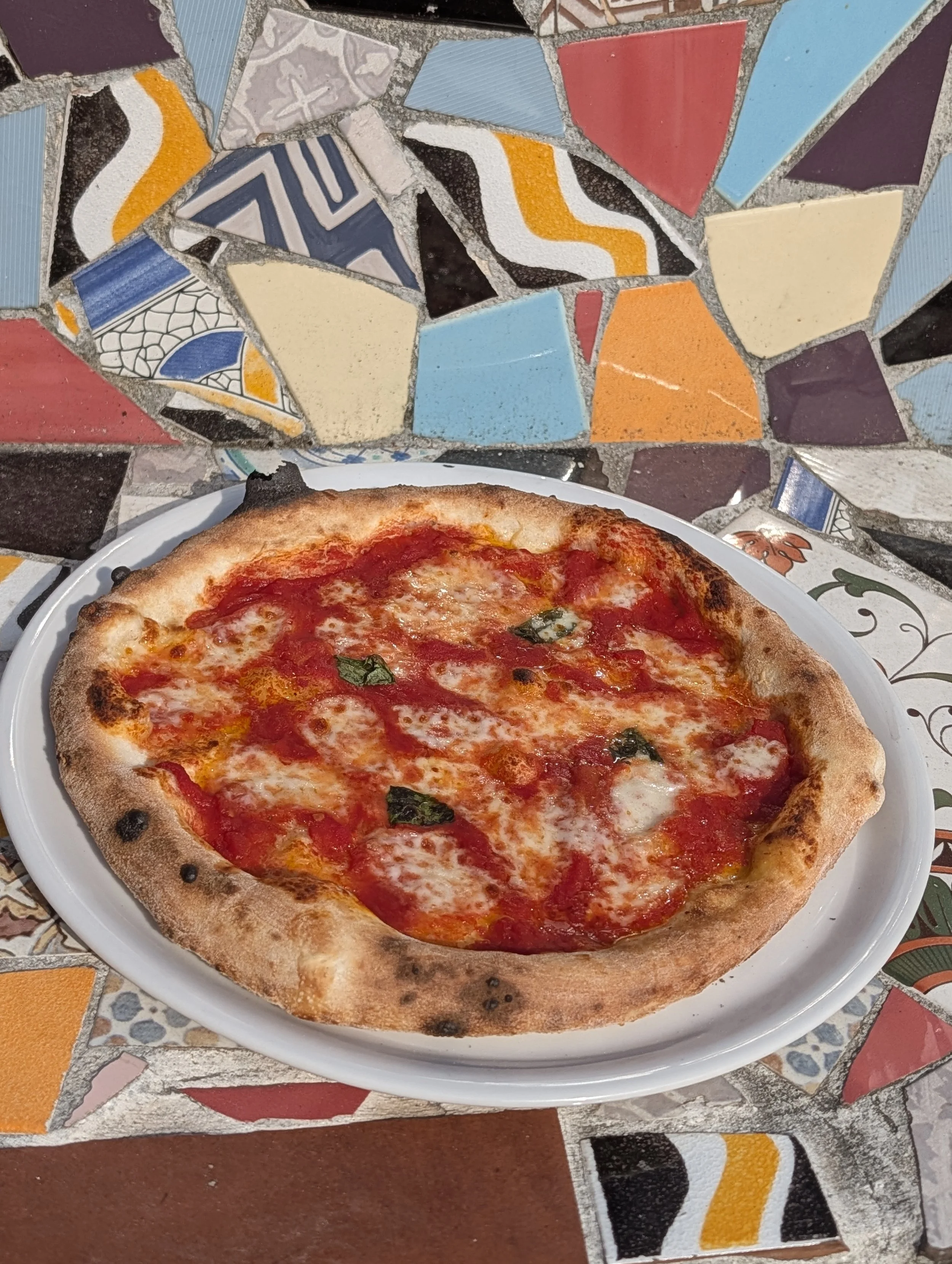 Eine Pizza mit Tomatensoße, Käse und Basilikum auf einem weißen Teller, auf einem bunten Mosaiktisch