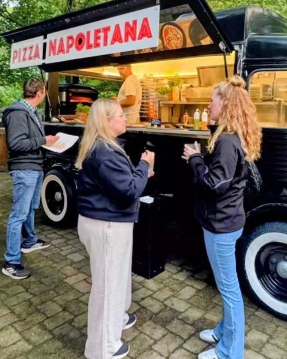 Der MEZZA Foodtruck mit der Aufschrift 'Pizza Napoletana' verkauft Pizza. Drei Gäste stehen vor dem Wagen und unterhalten sich beim Essen während der Pizzaiolo Pizza zubereitet.