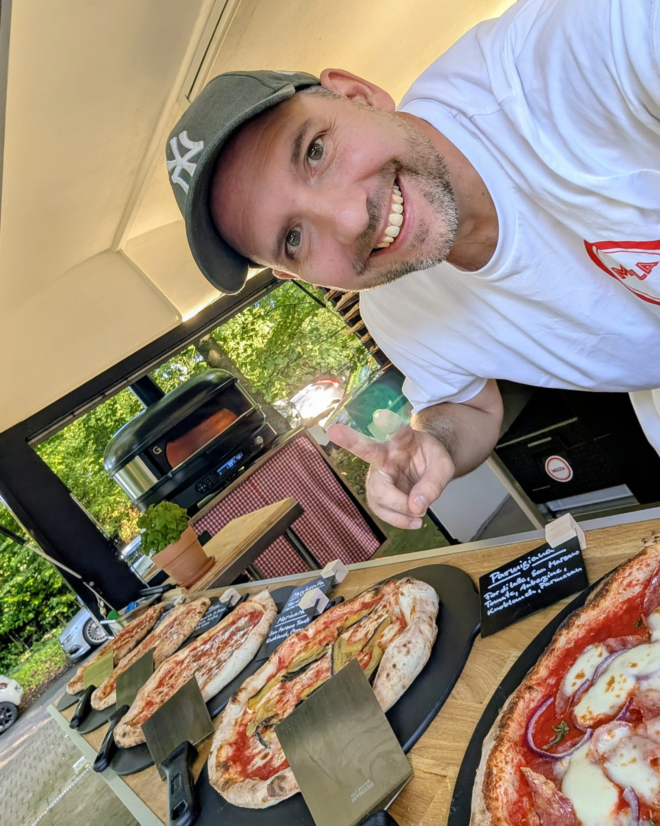 Ein Mann grinst in die Kamera und zeigt auf eine Auswahl an belegten Pizzastücken auf einem Tisch in einer Pizzeria. Im Hintergrund sind große Fenster mit Blick auf grüne Bäume.