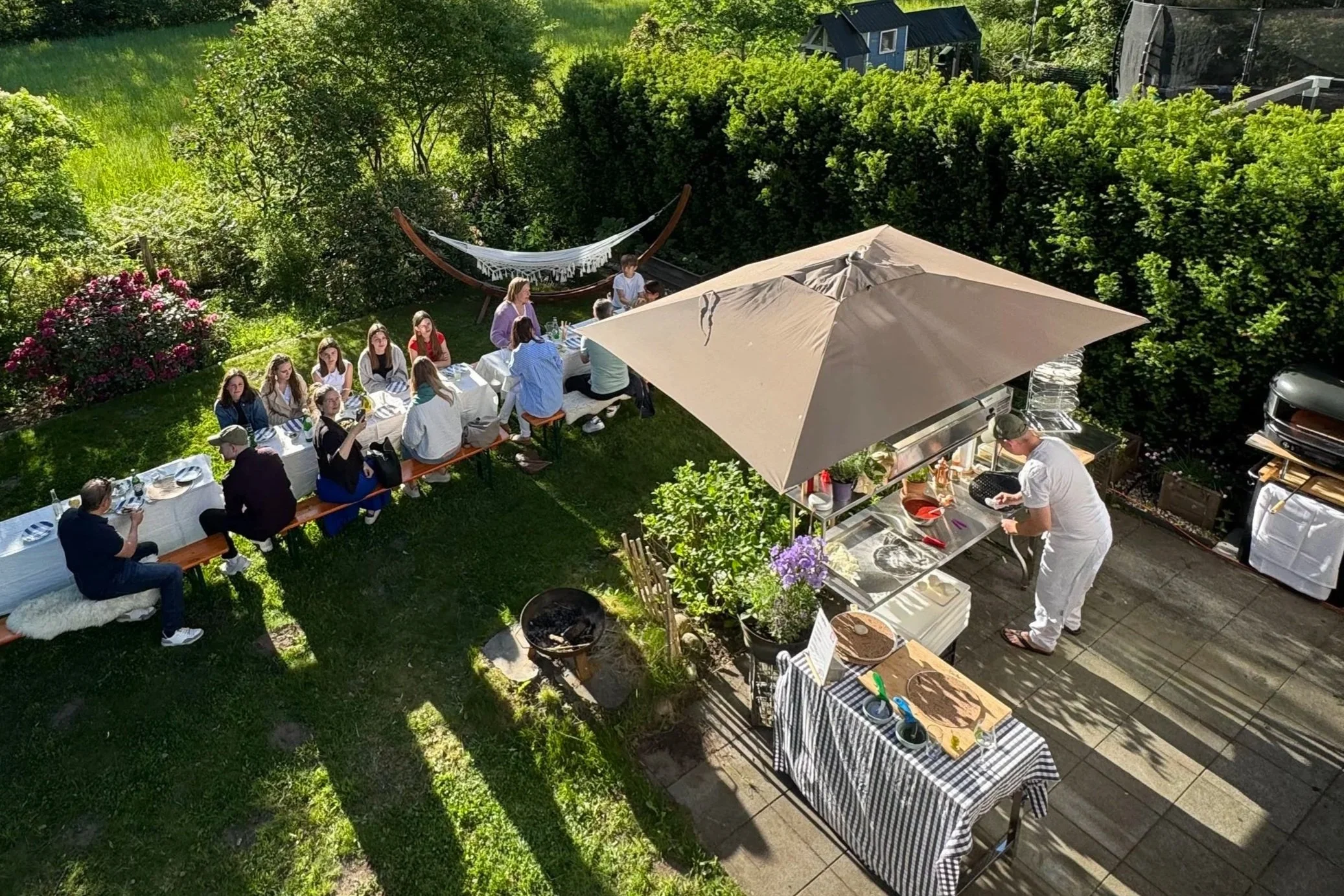 Eine Gartenparty im Grünen mit einem langen Esstisch, an dem Menschen sitzen, ein Mann, der am Grill arbeitet, und ein Mann, der eine Grillarbeit macht. Es gibt einen großen Sonnenschirm, Bäume und einen Trampolin im Hintergrund.