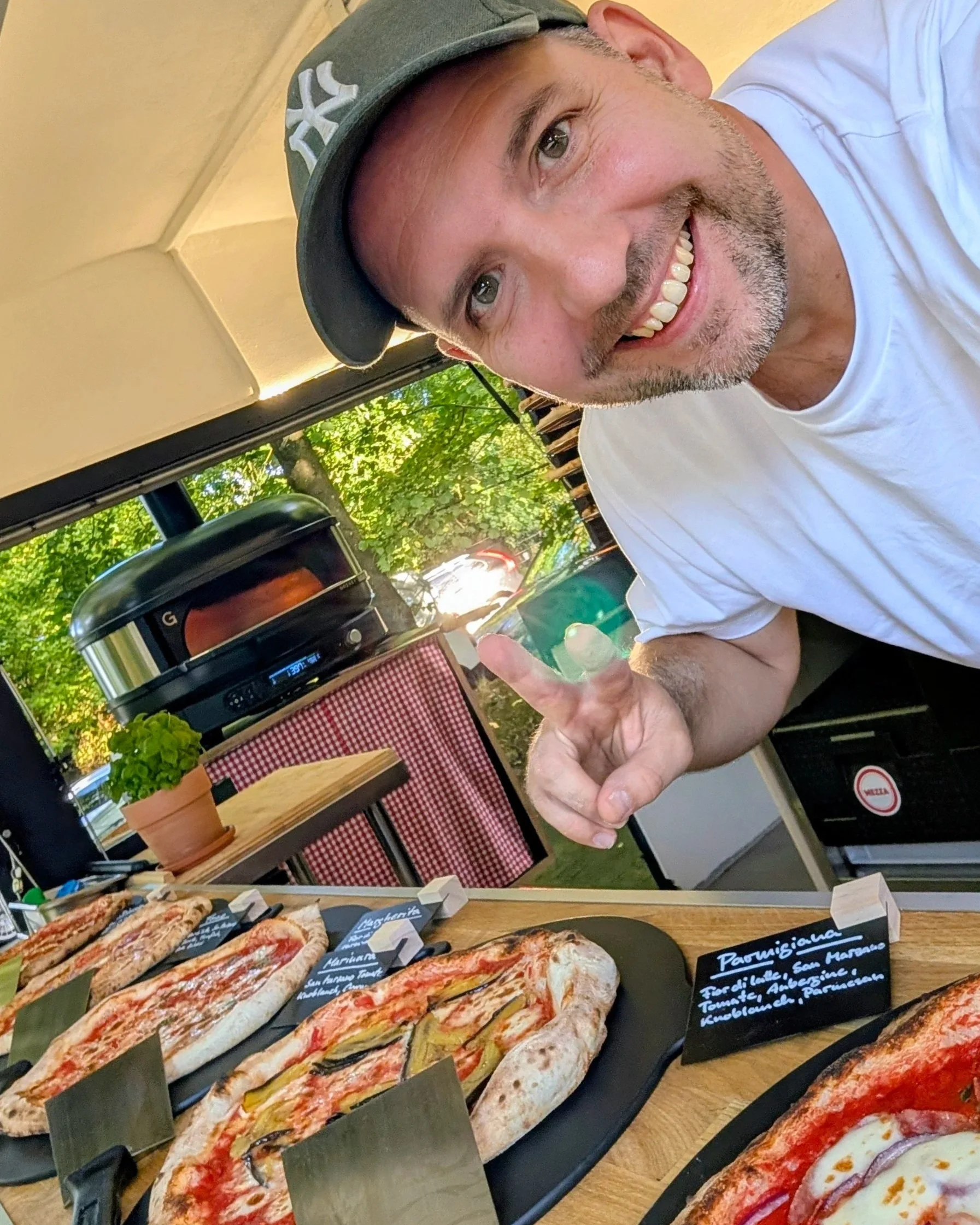Ein lächelnder Mann mit Kurzhaar und Bart zeigt auf verschiedene Pizzen auf einem Tisch. Im Hintergrund sind grüne Bäume und eine Terrasse sichtbar.