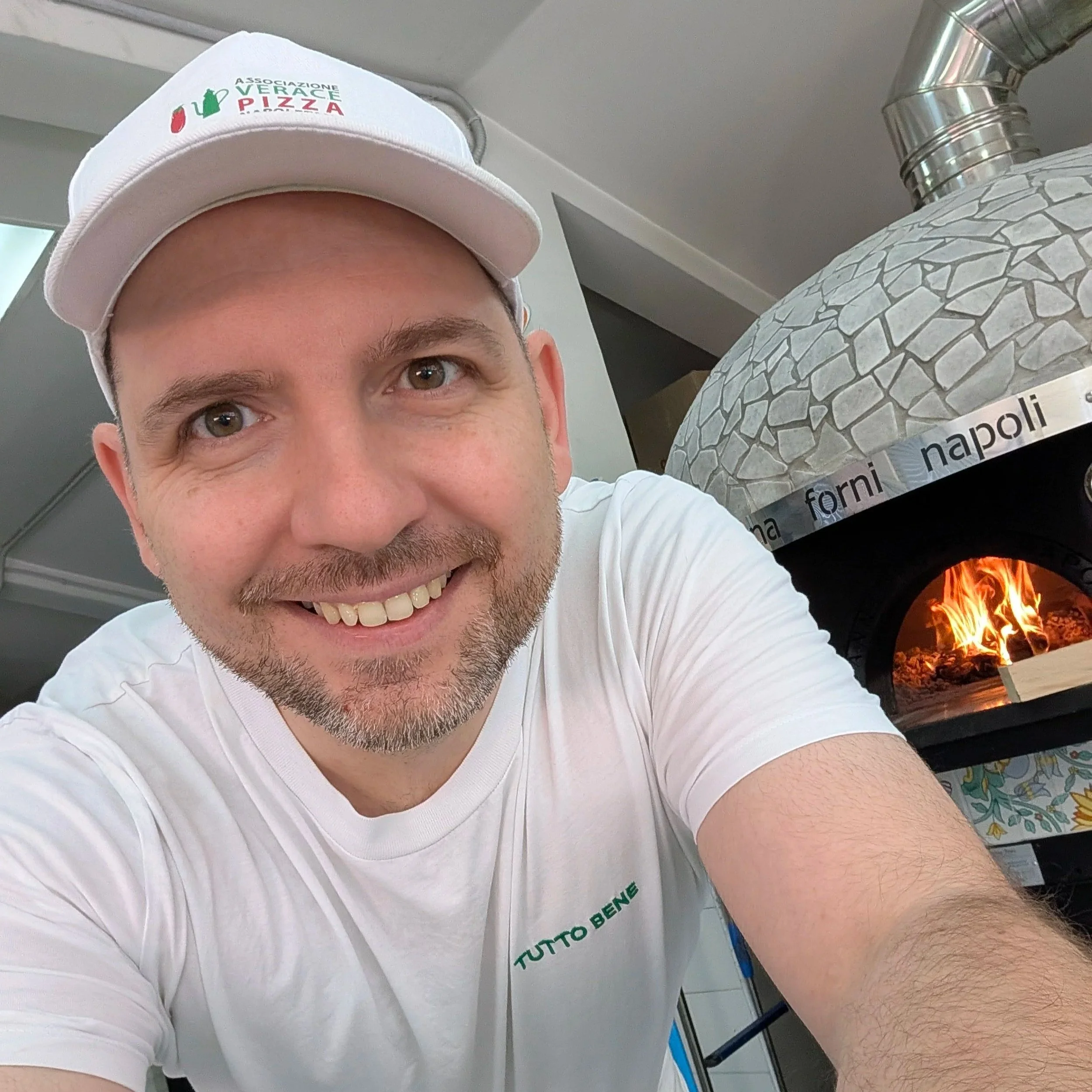 Ein junger Mann trägt ein weißes T-Shirt und eine weiße Mütze mit einem Logo und Text. Im Hintergrund ist ein Kamin mit Flammen sichtbar.