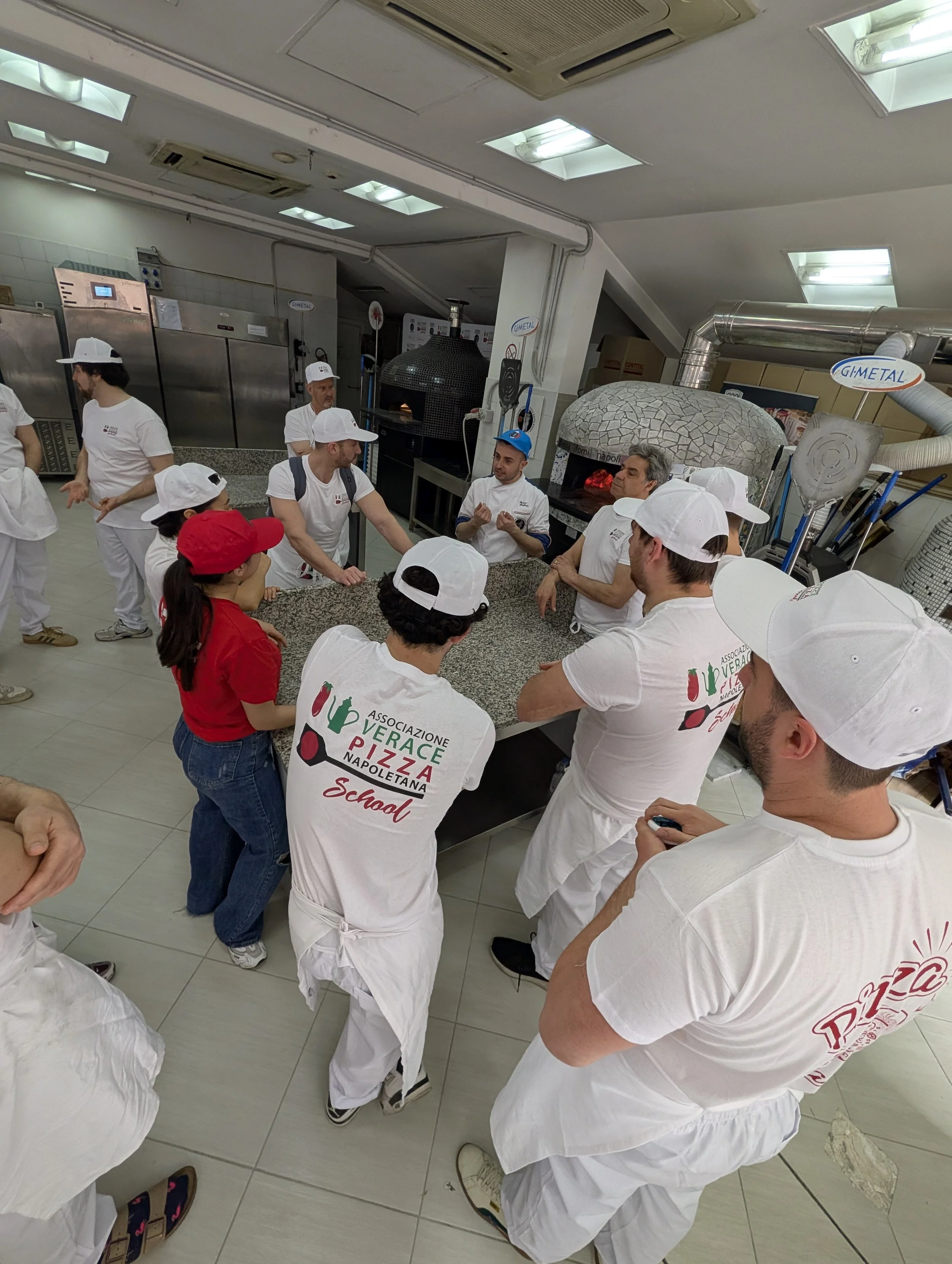 Mehrere Menschen in weißer Kochkleidung und weißen Kappen in einer Pizzeria, die anscheinend an einem Kochkurs oder einer Teambesprechung teilnehmen, vor eine Pizza-Backofen. Einige Personen diskutieren oder hören zu, während andere sich unterhalten.