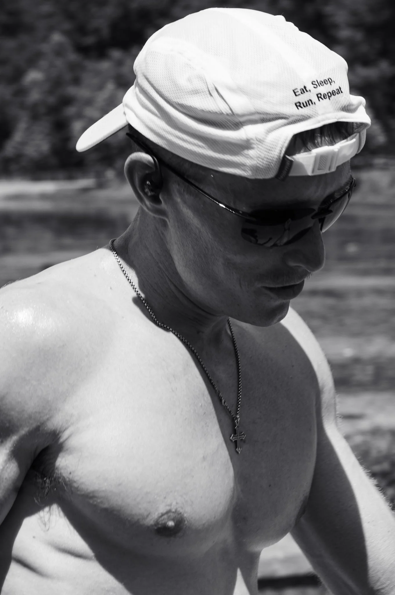 Close-up black and white photo of a man wearing a white cap with the words "Eat. Sleep. Run. Repeat." embroidered on it, sunglasses, a cross necklace, and a chain, at the beach.