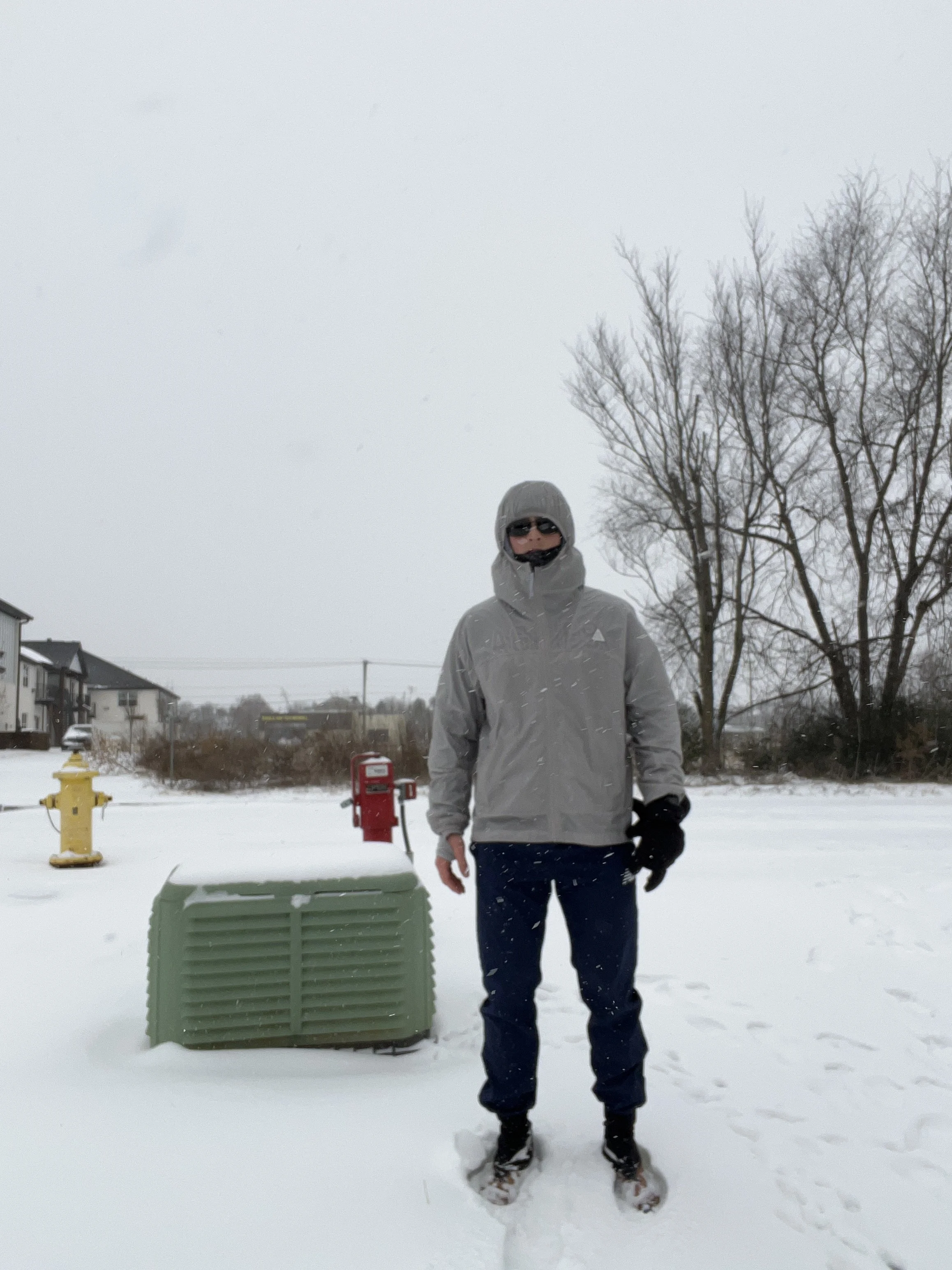 Person standing in snowy outdoor area wearing winter clothing and sunglasses, with trees and buildings in background.