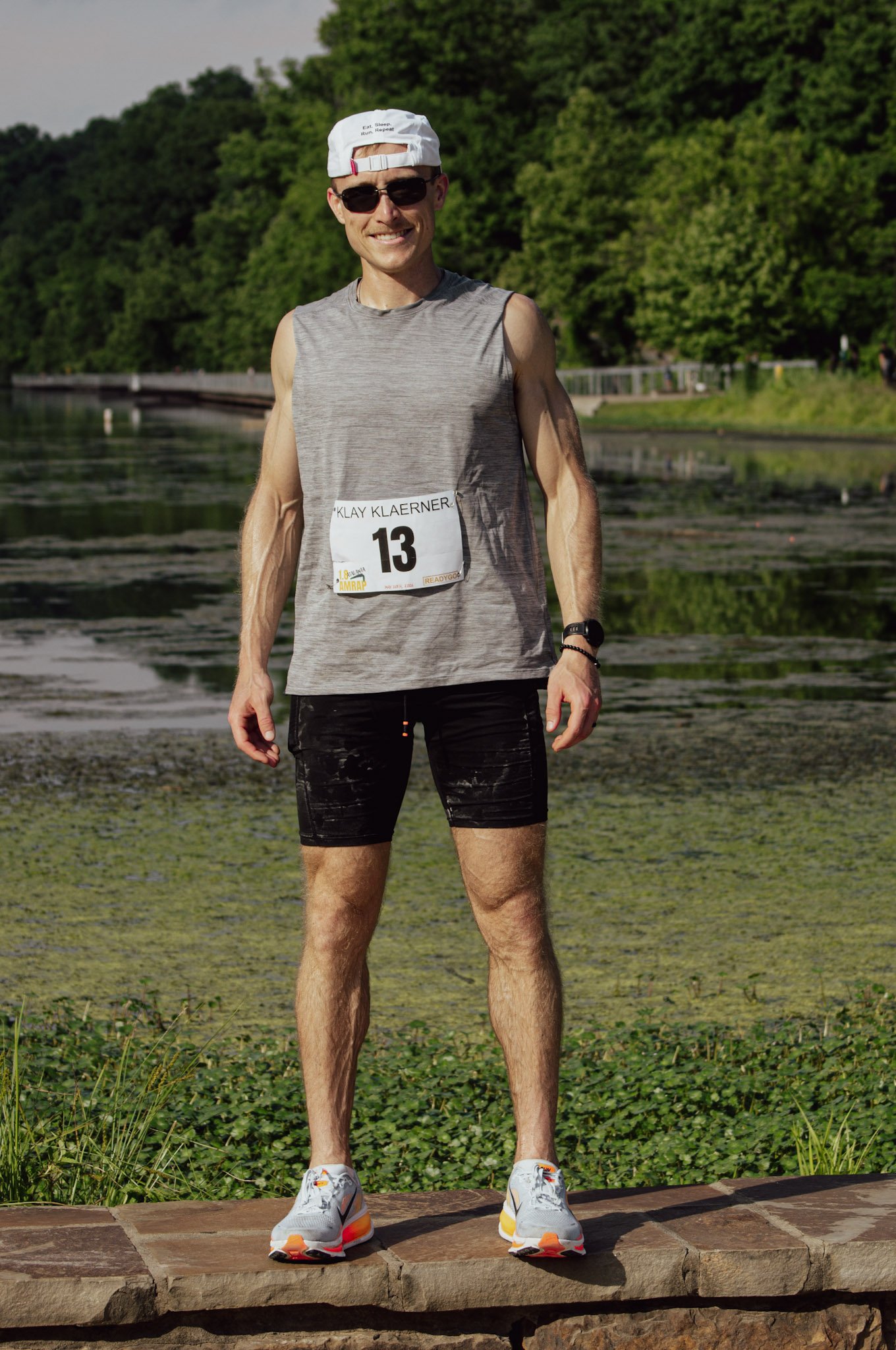 A smiling man in athletic attire standing outdoors on a sunny day, wearing a gray sleeveless shirt, black shorts, white sneakers with orange accents, sunglasses, a white cap backward, and a race number bib with the name 'Klay Klaerner' and the number 13. He is on a stone surface with greenery and trees in the background.
