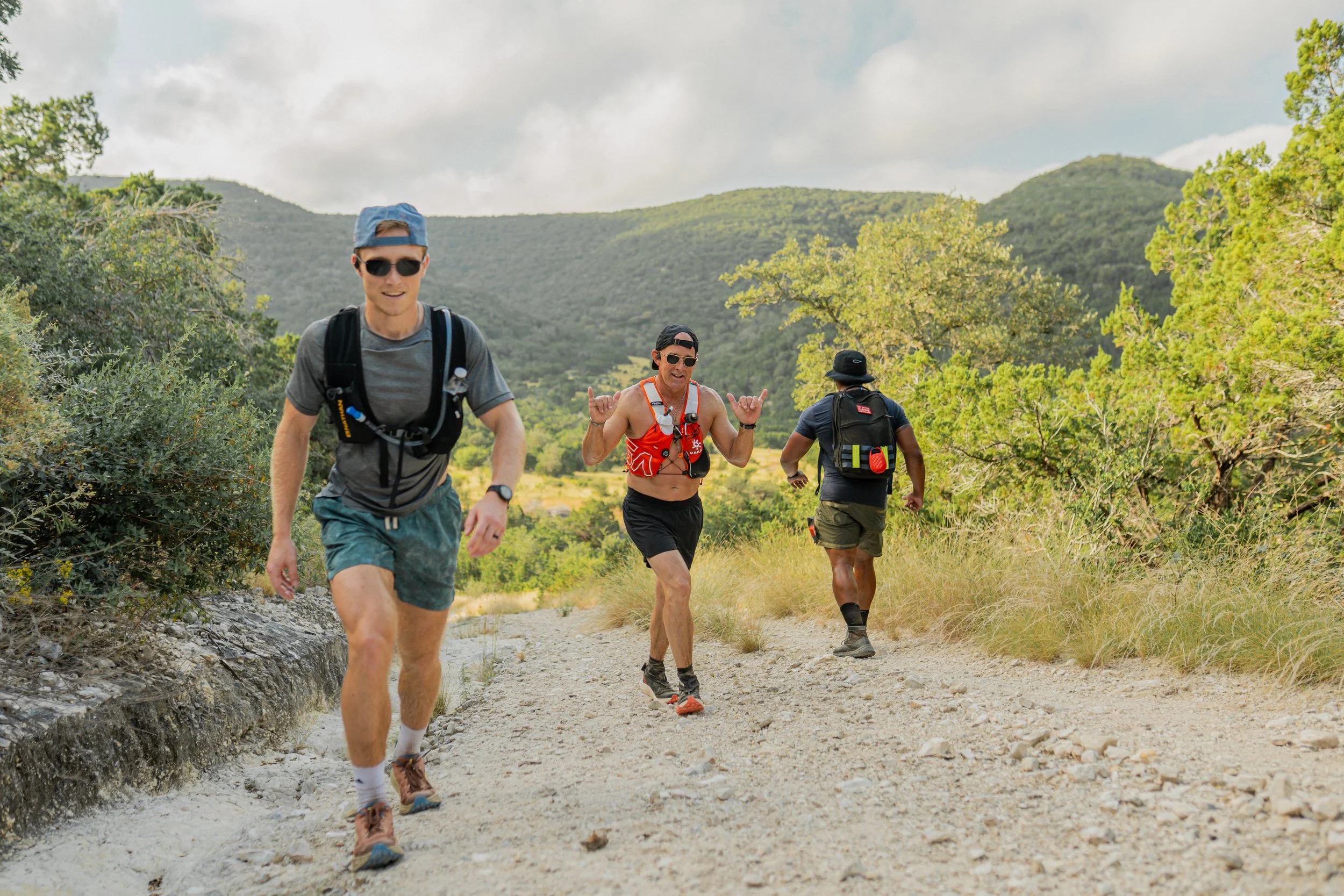 Four men trail running on a dirt trail in a natural setting with green trees and hills in the background, wearing athletic clothing and backpacks.