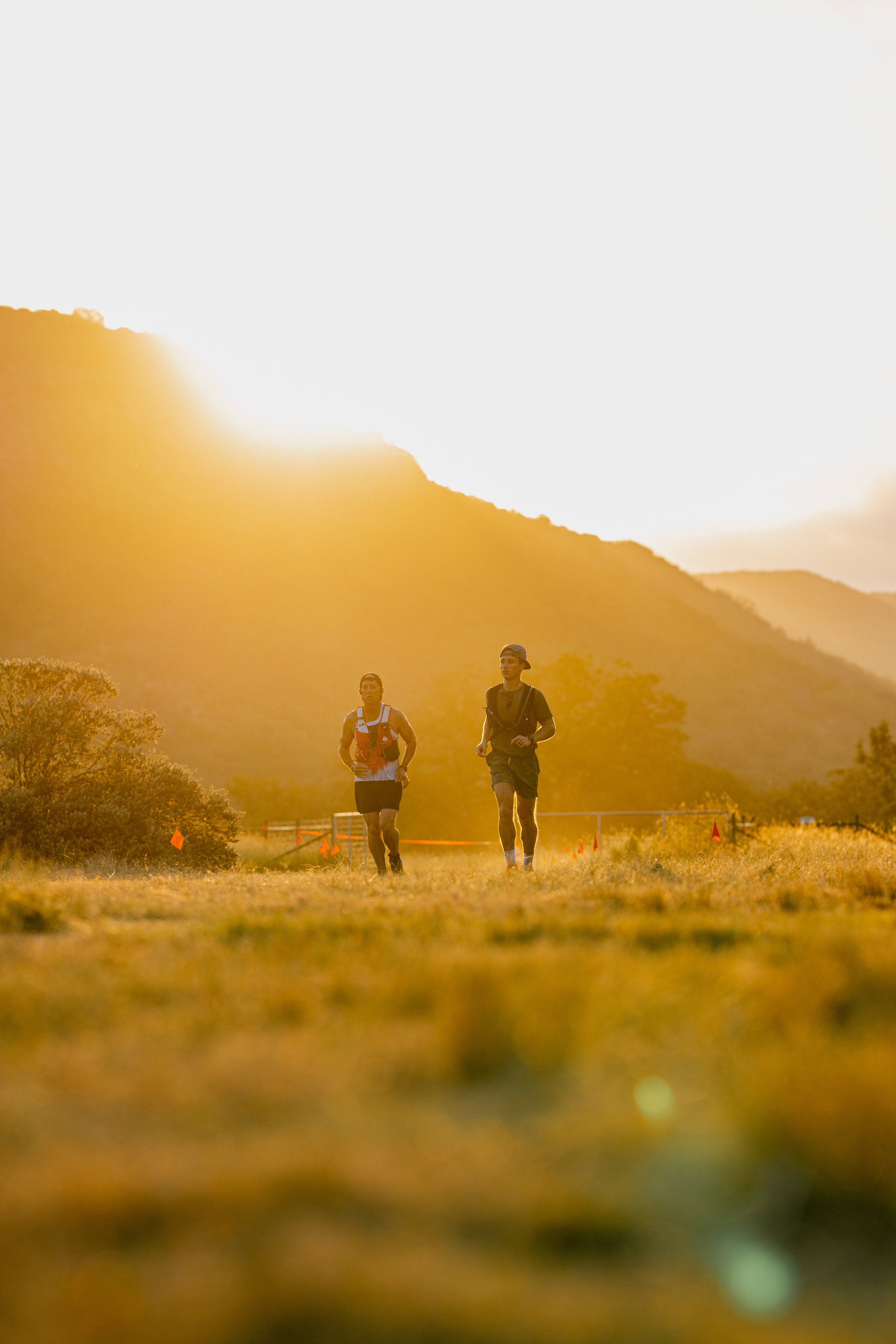 Two male runners participating in an outdoor race or marathon during sunset, with mountainous landscape in the background and orange safety cones along the trail.
