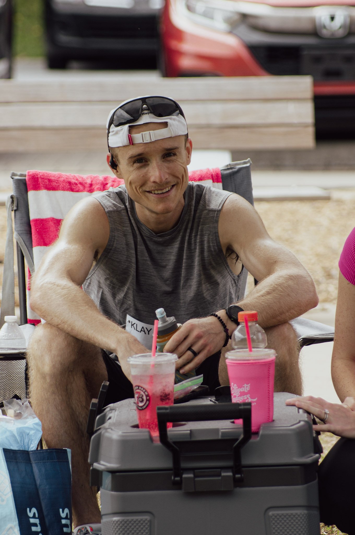 A smiling man wearing a white cap with sunglasses on top, seated at a table with drinks, surrounded by people and outdoor workout equipment.