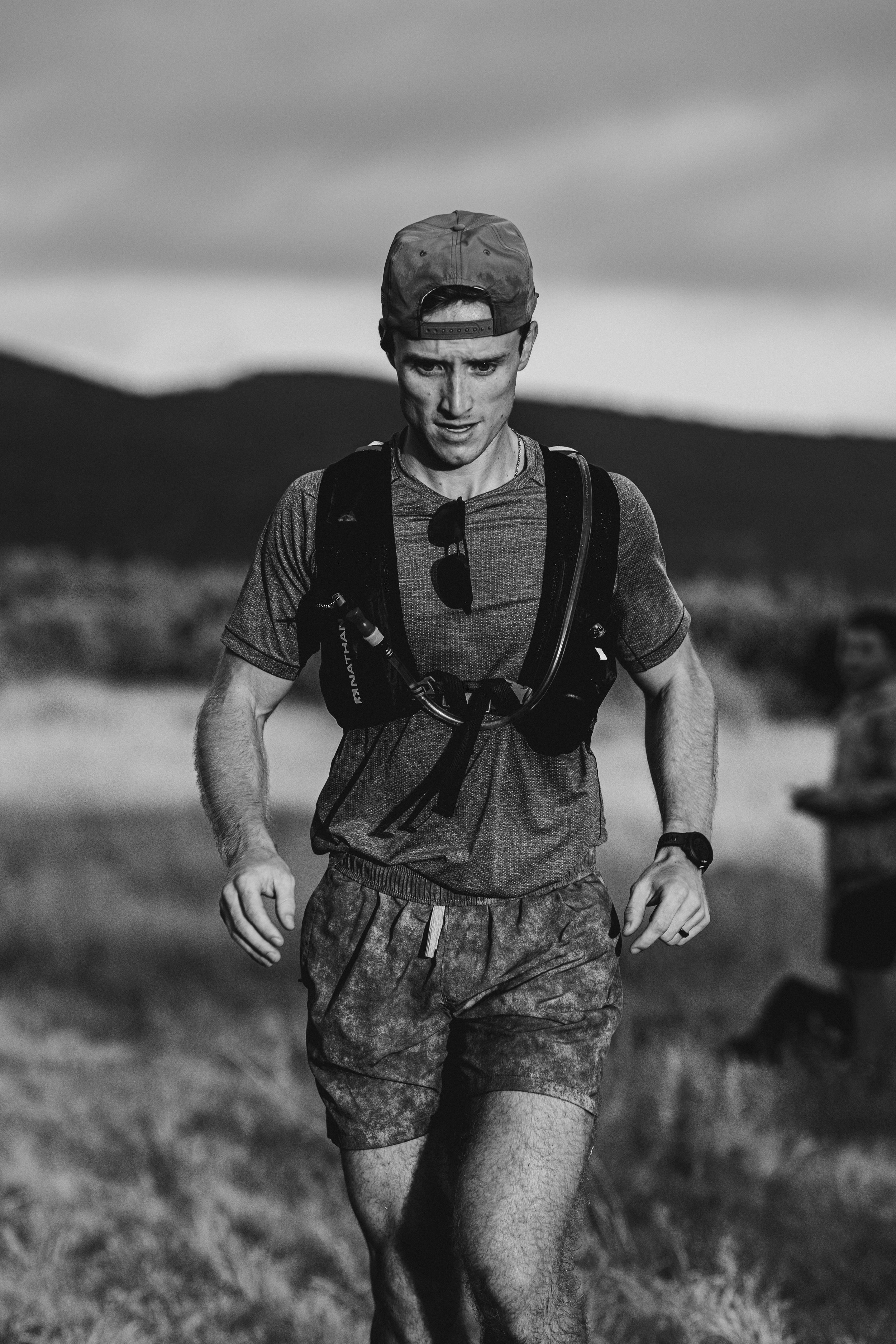 A young man running outdoors in a natural landscape, wearing a cap, t-shirt, shorts, and a hydration backpack.