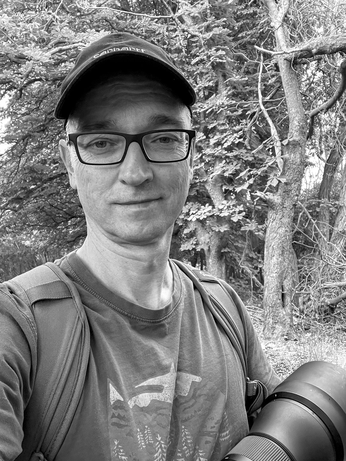 Ein Mann mit Brille, Baseballcap und Fotorucksack steht in einem Wald und hält eine Kamera in der Hand.