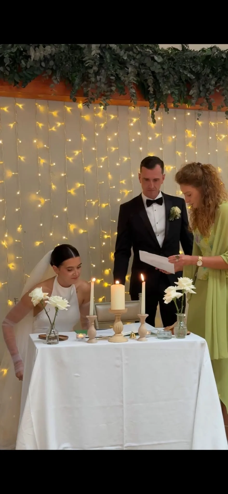 A wedding ceremony with a bride, groom, and officiant standing behind a table decorated with candles and flowers, against a backdrop of string lights and greenery.