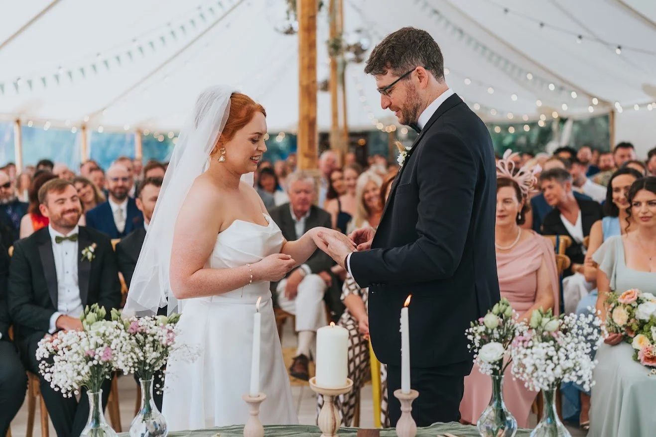 A bride and groom exchanging rings during their wedding ceremony inside a decorated tent with guests seated in the background.