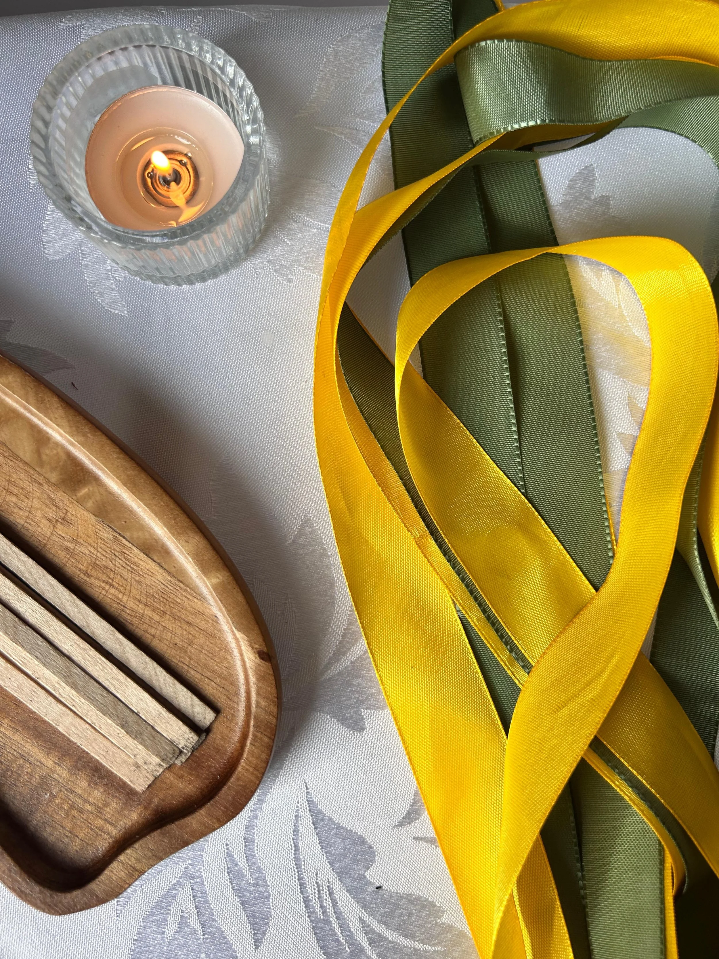 A lit candle in a glass holder, a wooden spatula, and yellow and green ribbon on a white tablecloth with floral pattern.