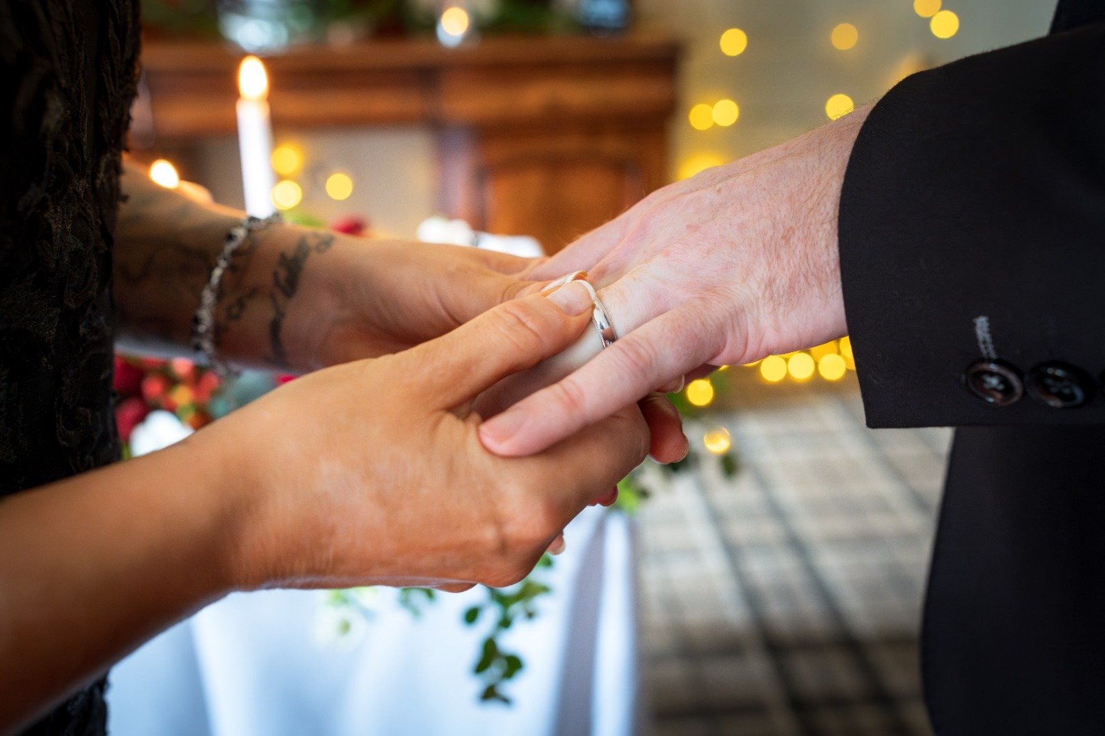 Close-up of a couple exchanging wedding rings during a wedding ceremony, with blurred background of candles and decorations.