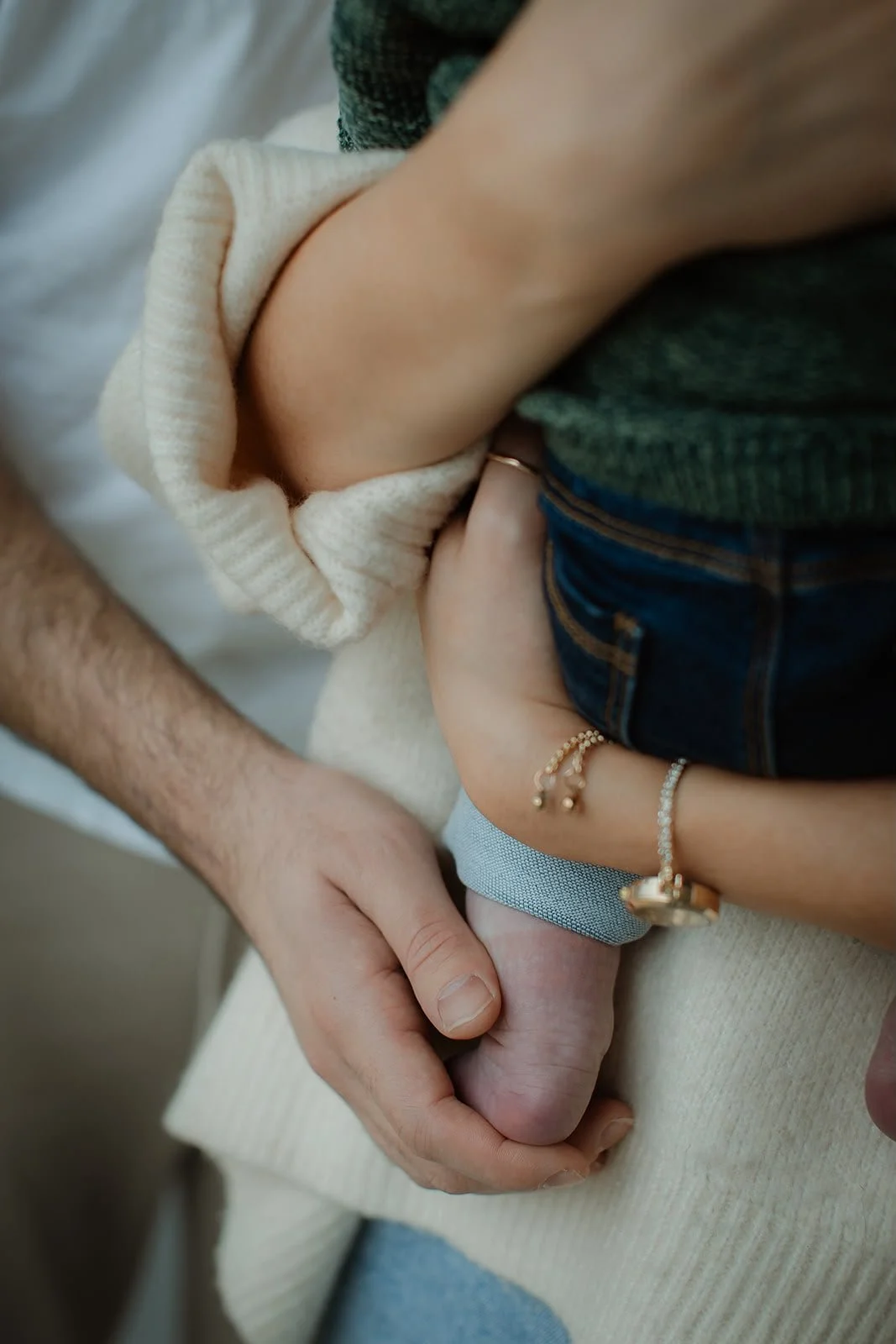 Close-up of two people holding hands, with one person wearing a beige knit sweater and the other wearing blue jeans, rings, and bracelets.