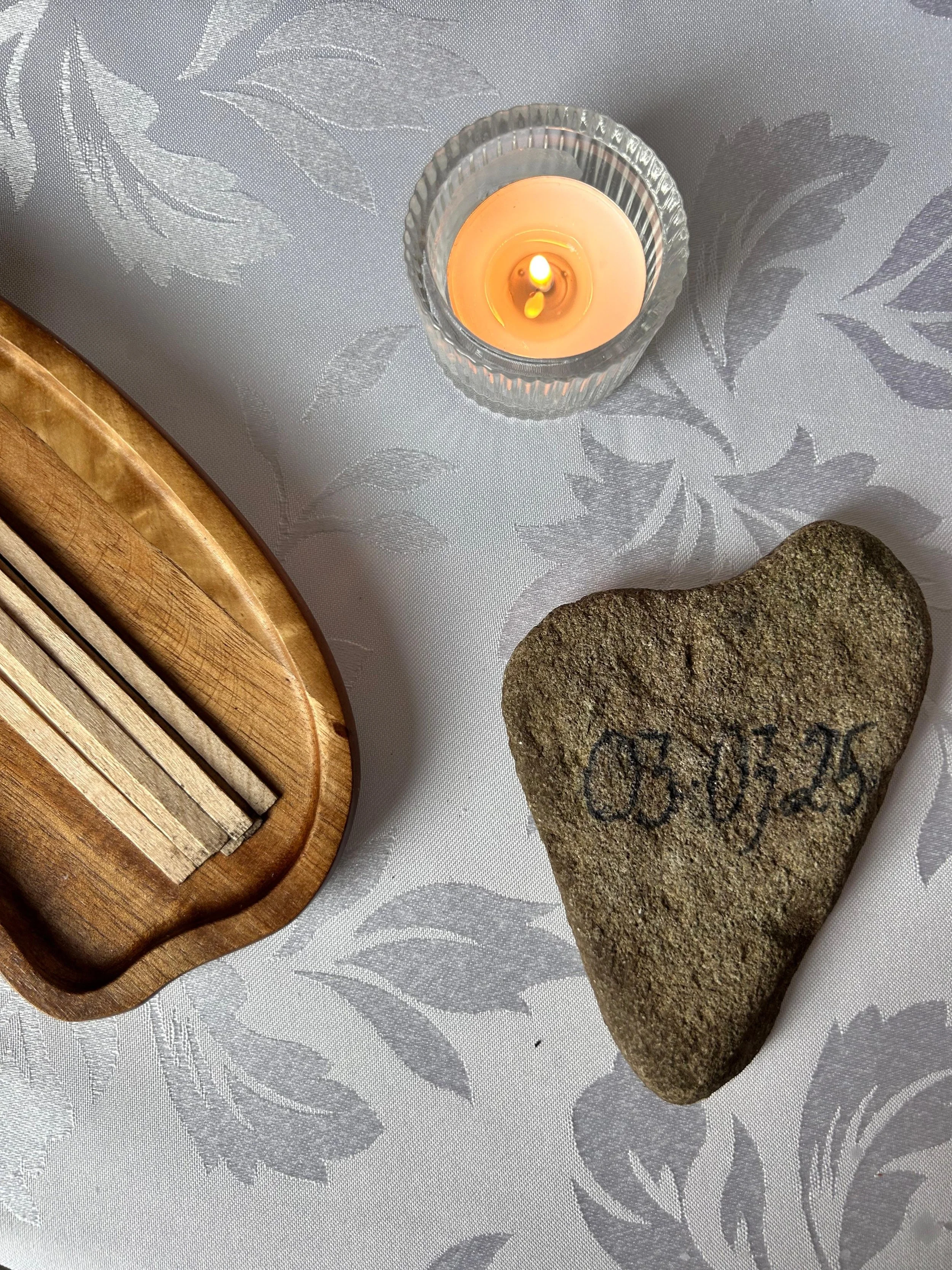 A lit candle in a glass holder, a heart-shaped stone with the word "LOVE" engraved on it, and a wooden tray on a table with a white floral tablecloth.