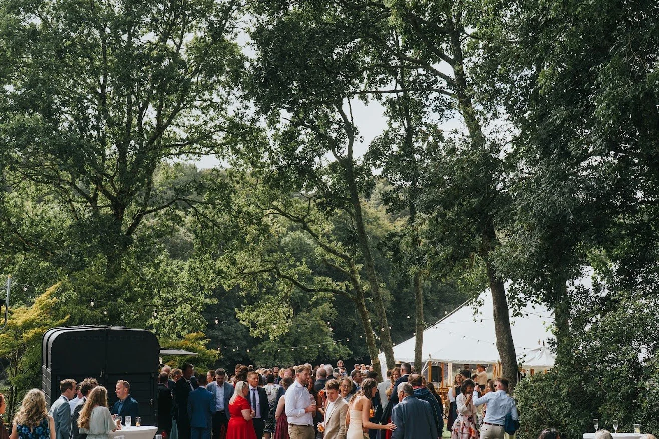 People attending an outdoor event or wedding reception under a large white tent, surrounded by green trees, with a string of lights overhead.