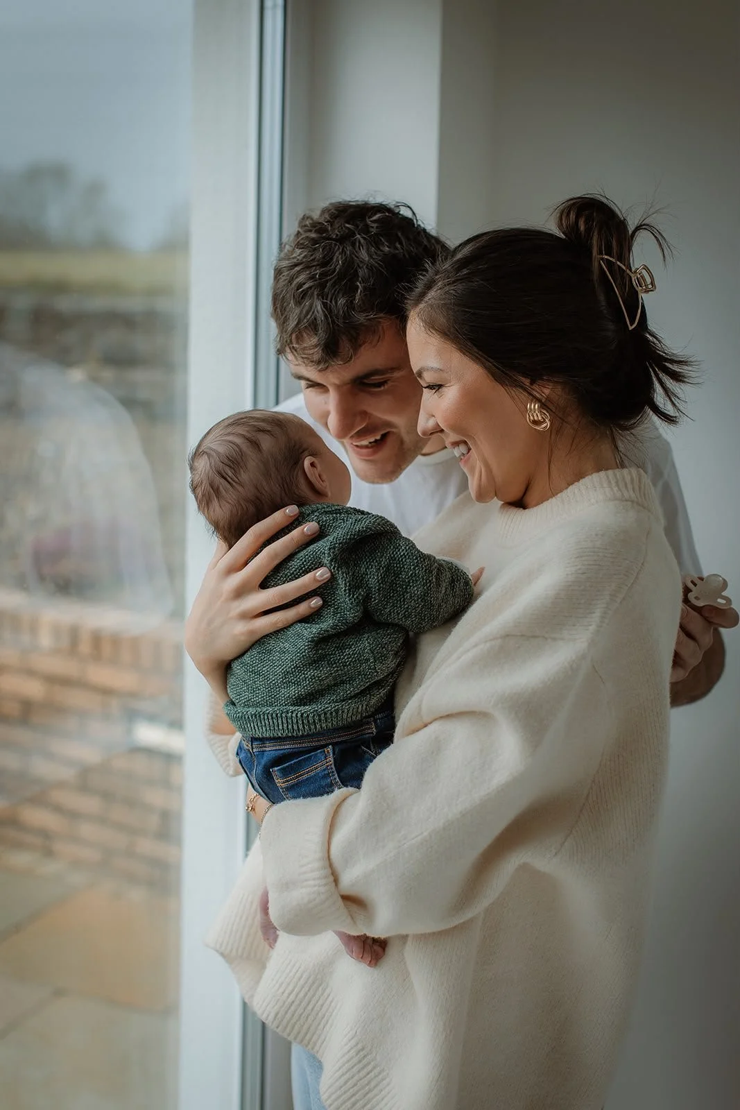 A happy family of three, a man, a woman, and a baby, standing by a window and smiling at each other warmly.