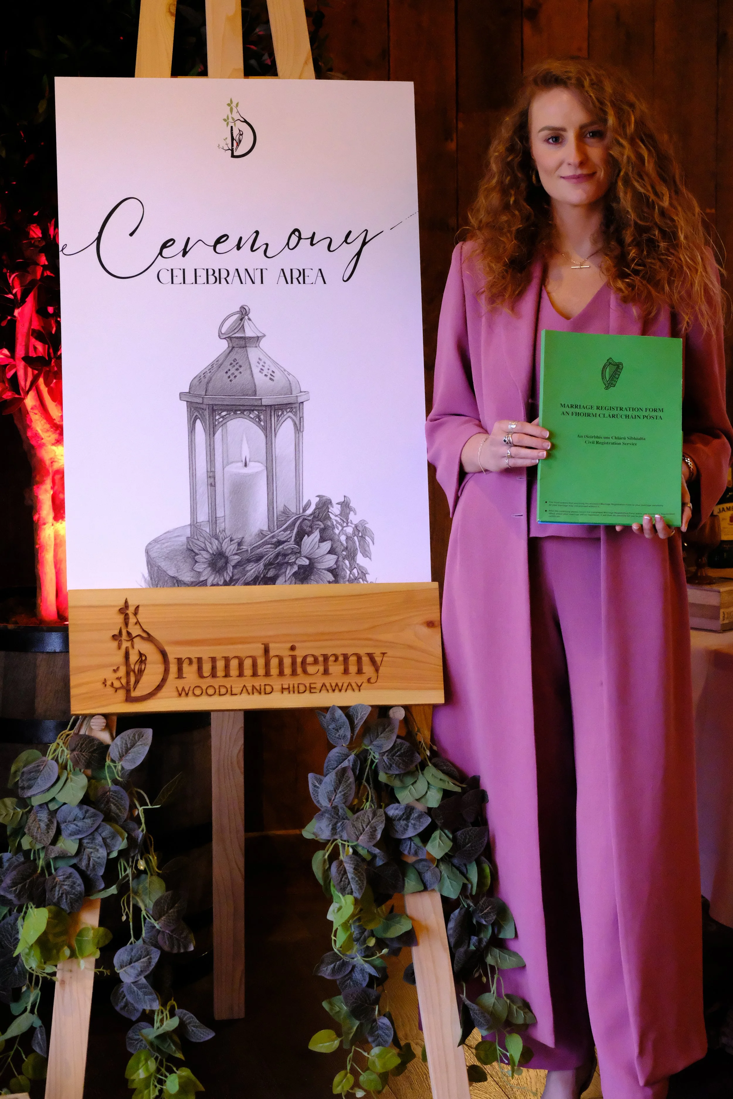 A woman with curly red hair wearing a pink outfit holding a green marriage registration form. Behind her an easel with a white sign reading "Ceremony Celebrant Area" with a drawing of a wedding lantern and flowers. There are plants at the bottom of the image and wooden walls in the background.
