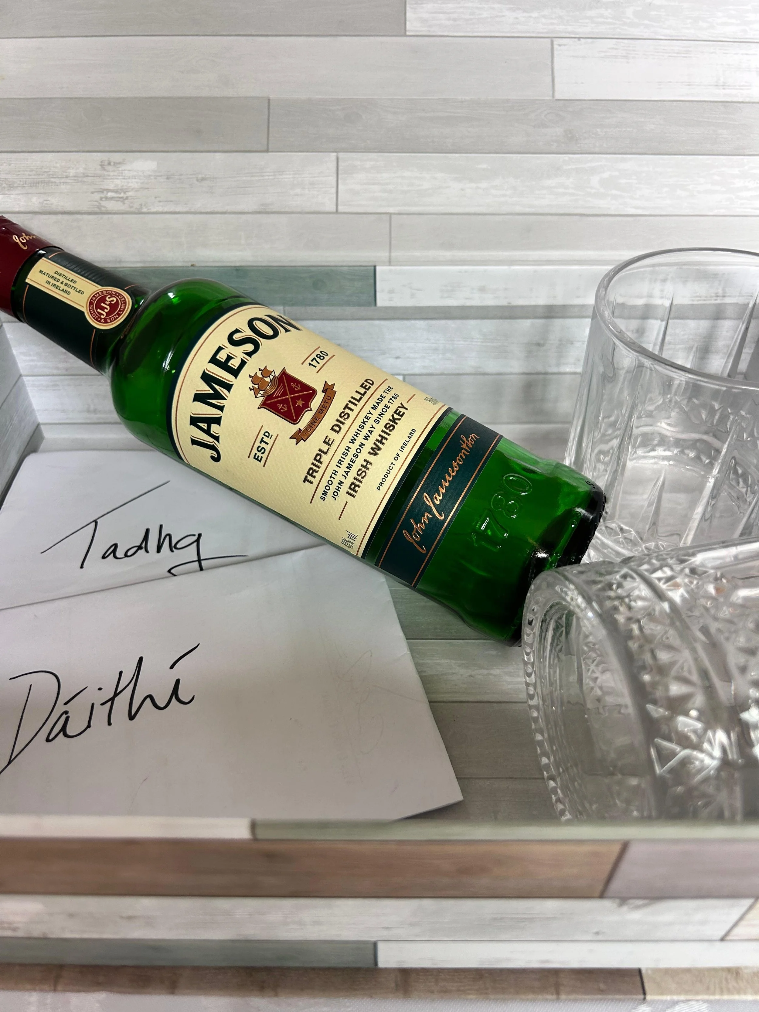 A green bottle of Jameson Irish whiskey lying on its side on a table. Next to the bottle are three clear, patterned glasses. A piece of paper with handwritten text is partially visible beneath the bottle.