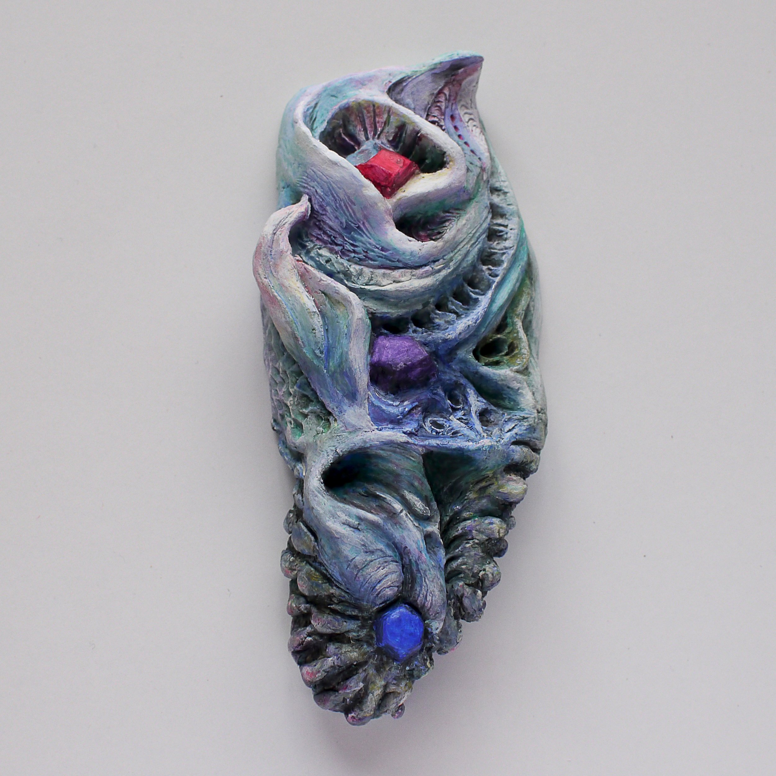 A colorful, abstract sculpture resembling a distorted face or skull with a red rectangular element inside the open mouth, set against a plain white background.