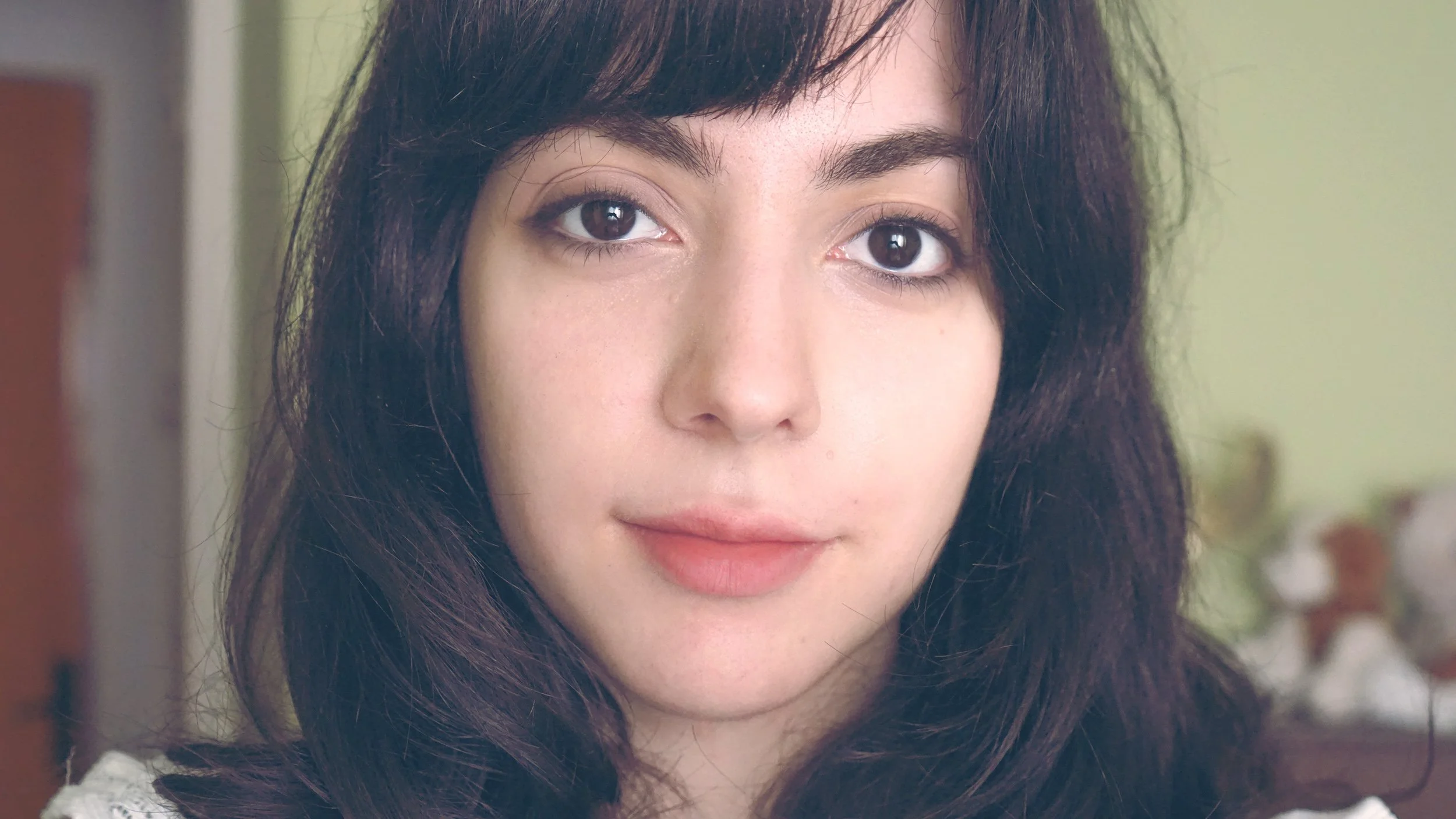 Close-up of a young woman's face with long dark hair, brown eyes, and a gentle smile, indoors with a blurred background.