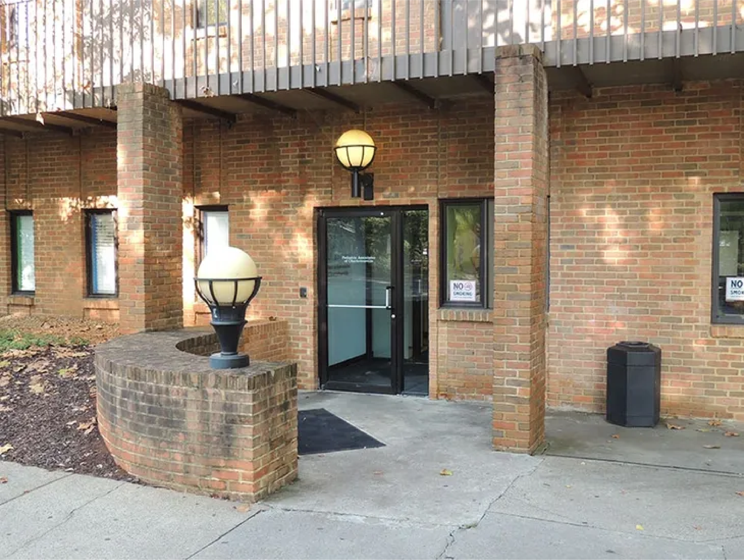 Front view of a brick building with a door and several windows, two decorative lamps, and a small brick wall with a curved corner at the entrance, surrounded by pavement.