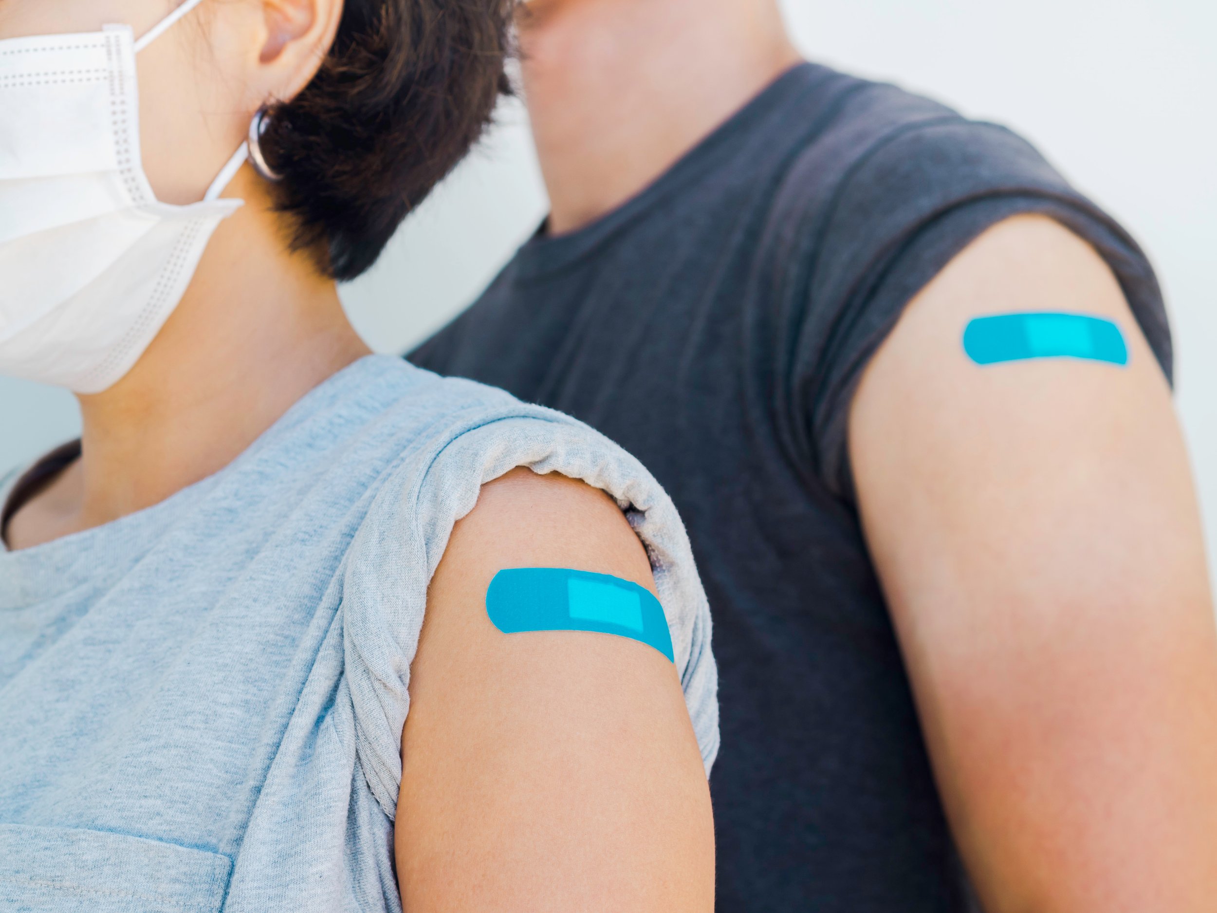 A person with a beard wearing a gray shirt and a face mask, receiving a vaccine shot in their upper arm, with blue medical tape applied over the injection site.