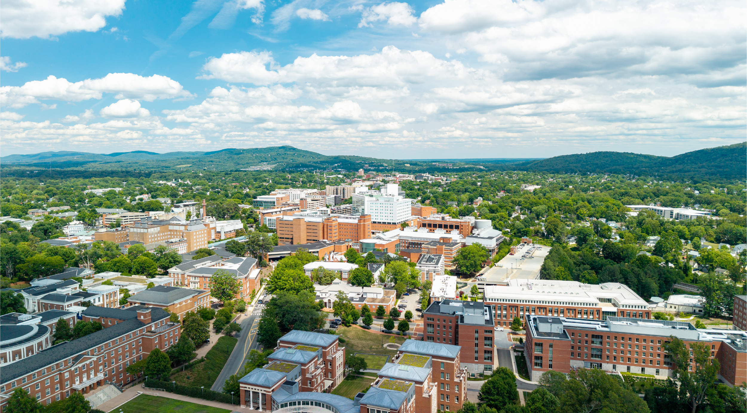 Aerial view of a city with a mix of brick and modern buildings surrounded by green trees, with hills and a partly cloudy sky in the background.