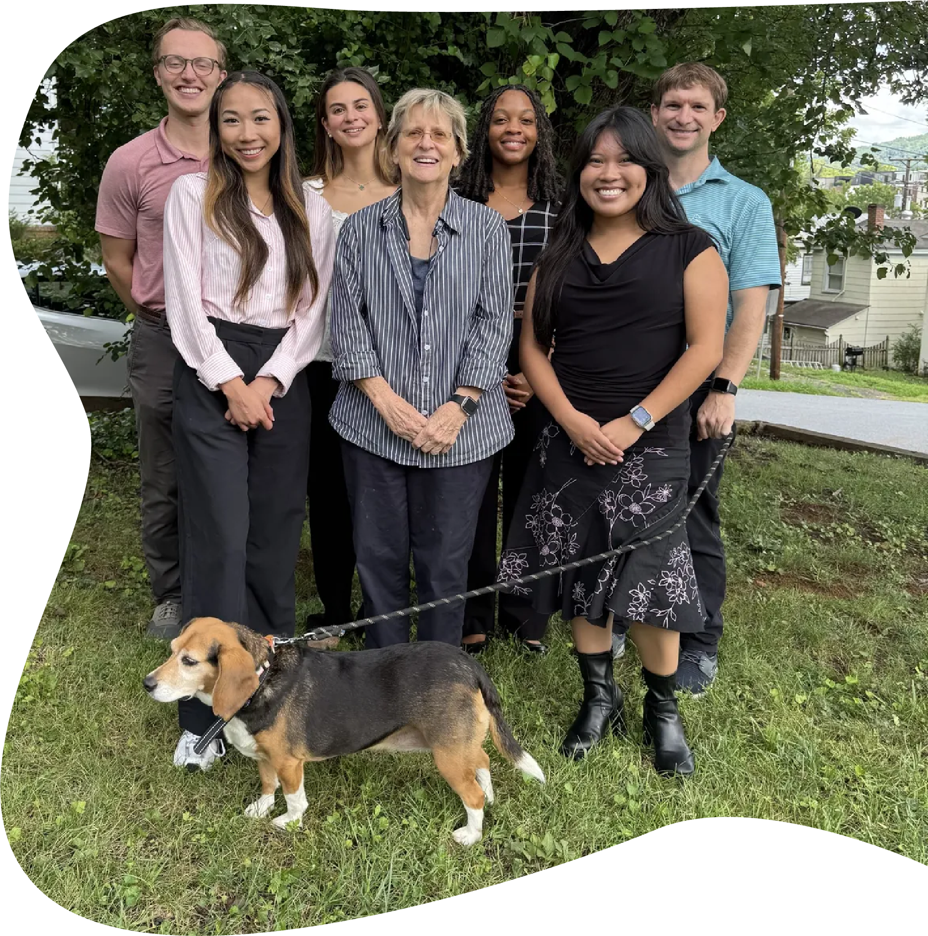 A group of eight diverse people standing outdoors on grass in front of trees, with one dog in front. The group includes young adults, middle-aged, and an older woman, all smiling and dressed casually. The dog is a beagle with black, brown, and white fur.
