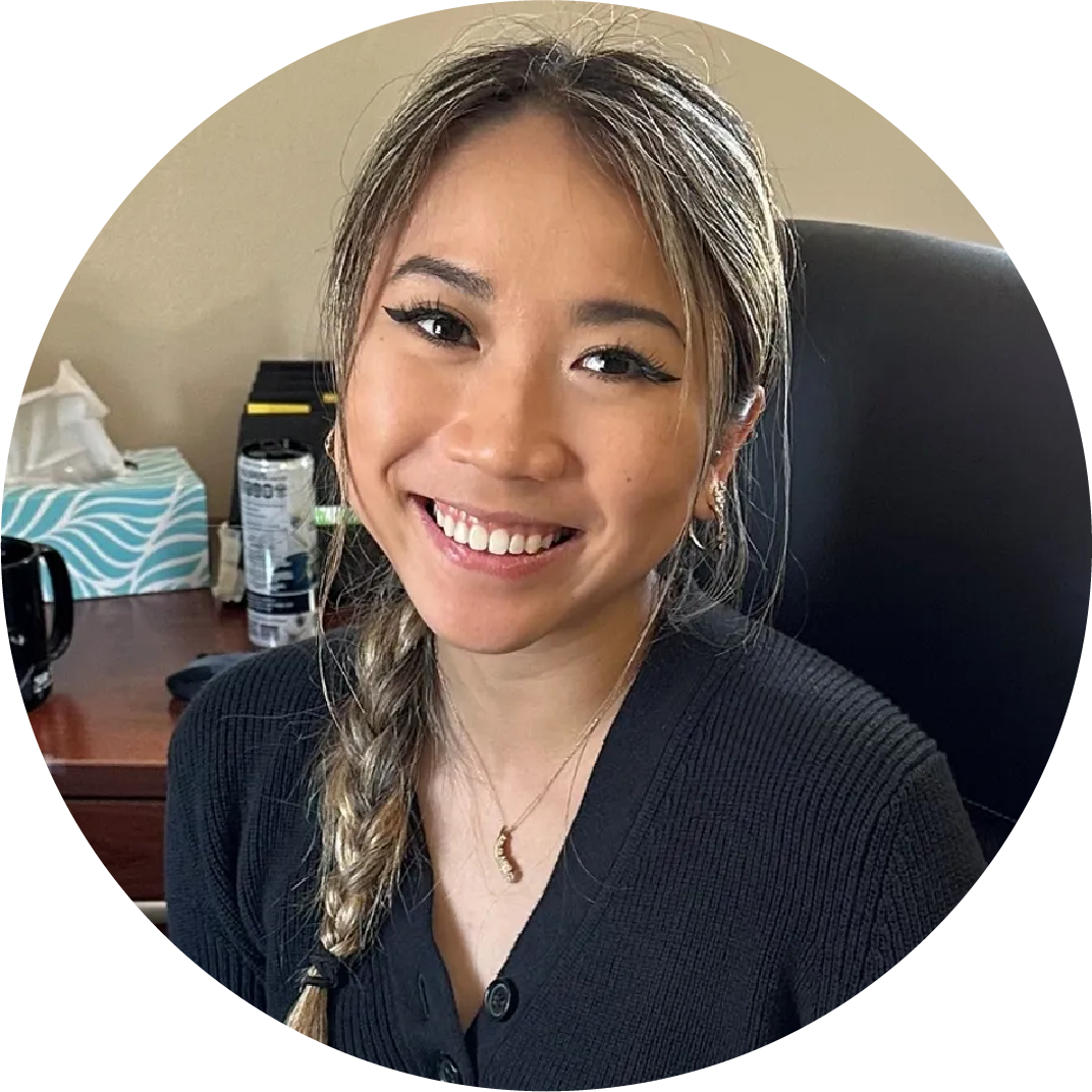 A smiling woman, Catherine Hansa, with long hair in a braid, wearing a black shirt and necklace, sitting at a desk.