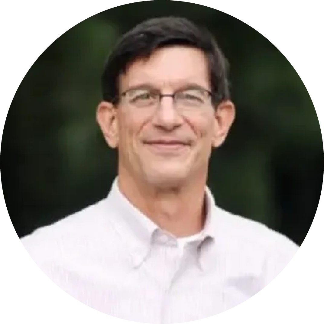 Portrait of a man, Paul Wisman Jr., with dark hair, glasses, and wearing a white collared shirt, smiling outdoors against a blurred green background.