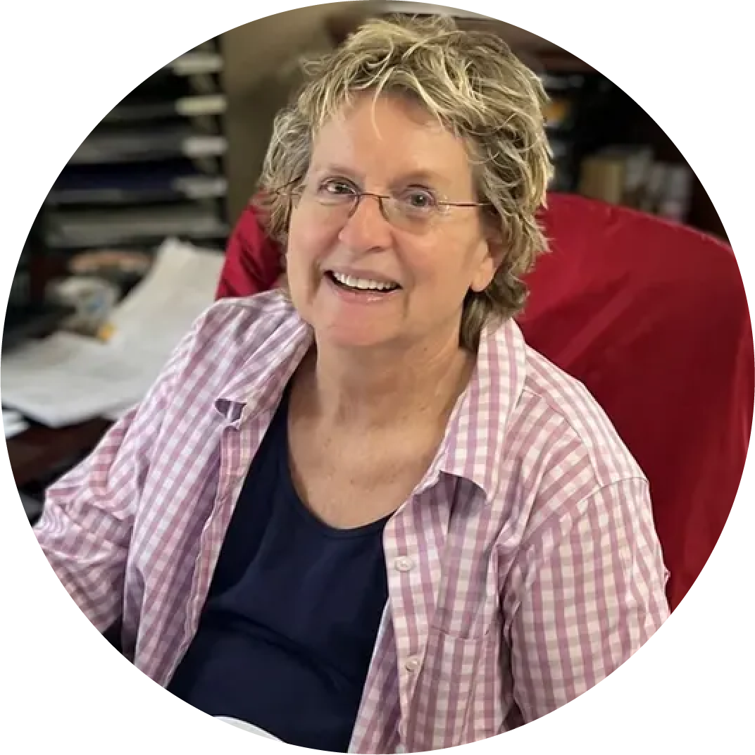 A smiling woman, Poody Wisman, with curly, short blonde hair and glasses sitting at a desk in an office.
