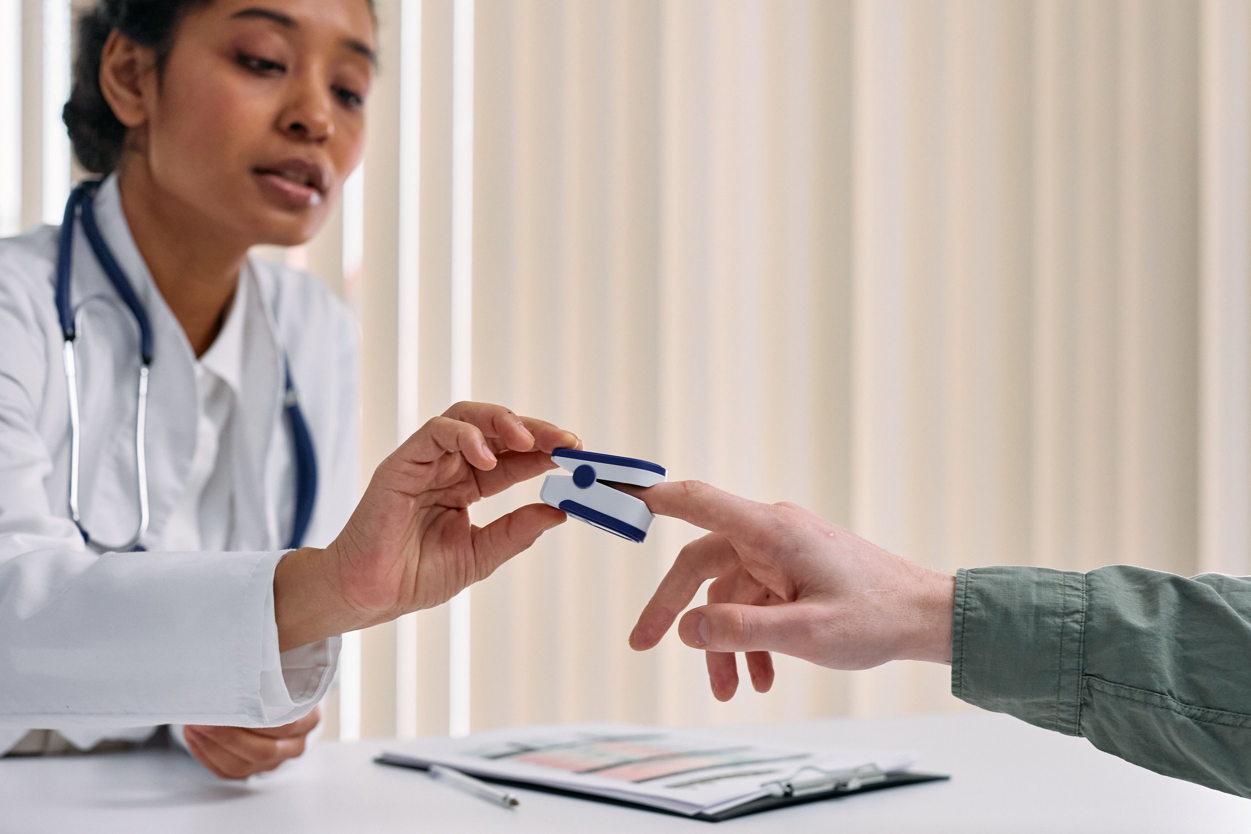 A healthcare professional, likely a doctor, is handing a blood glucose meter to a patient. The doctor is wearing a white coat and has a stethoscope around her neck. The scene appears to take place in a medical setting with a table, clipboard, and pen in the background.