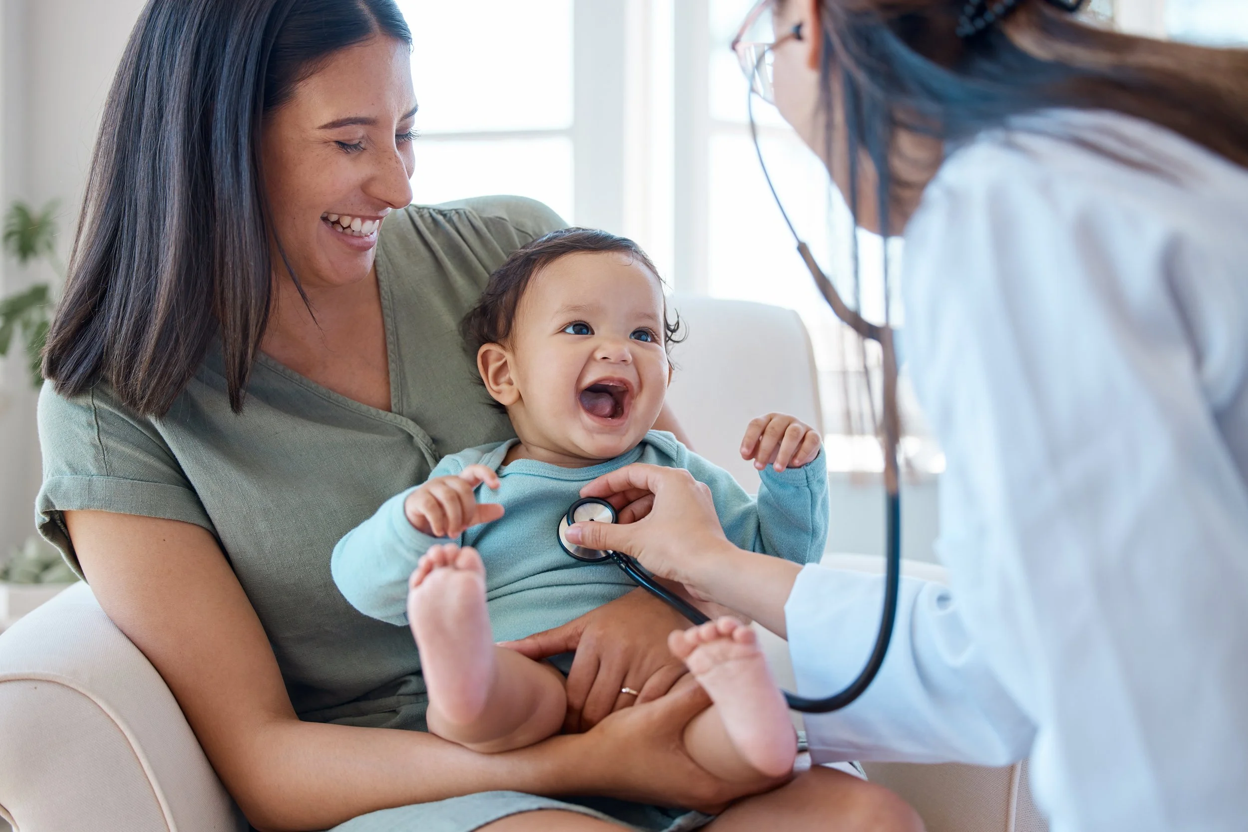 A smiling woman sitting on a couch holding a young child, while a medical professional with a stethoscope examines the child. The scene takes place indoors near a window, with natural light coming in.