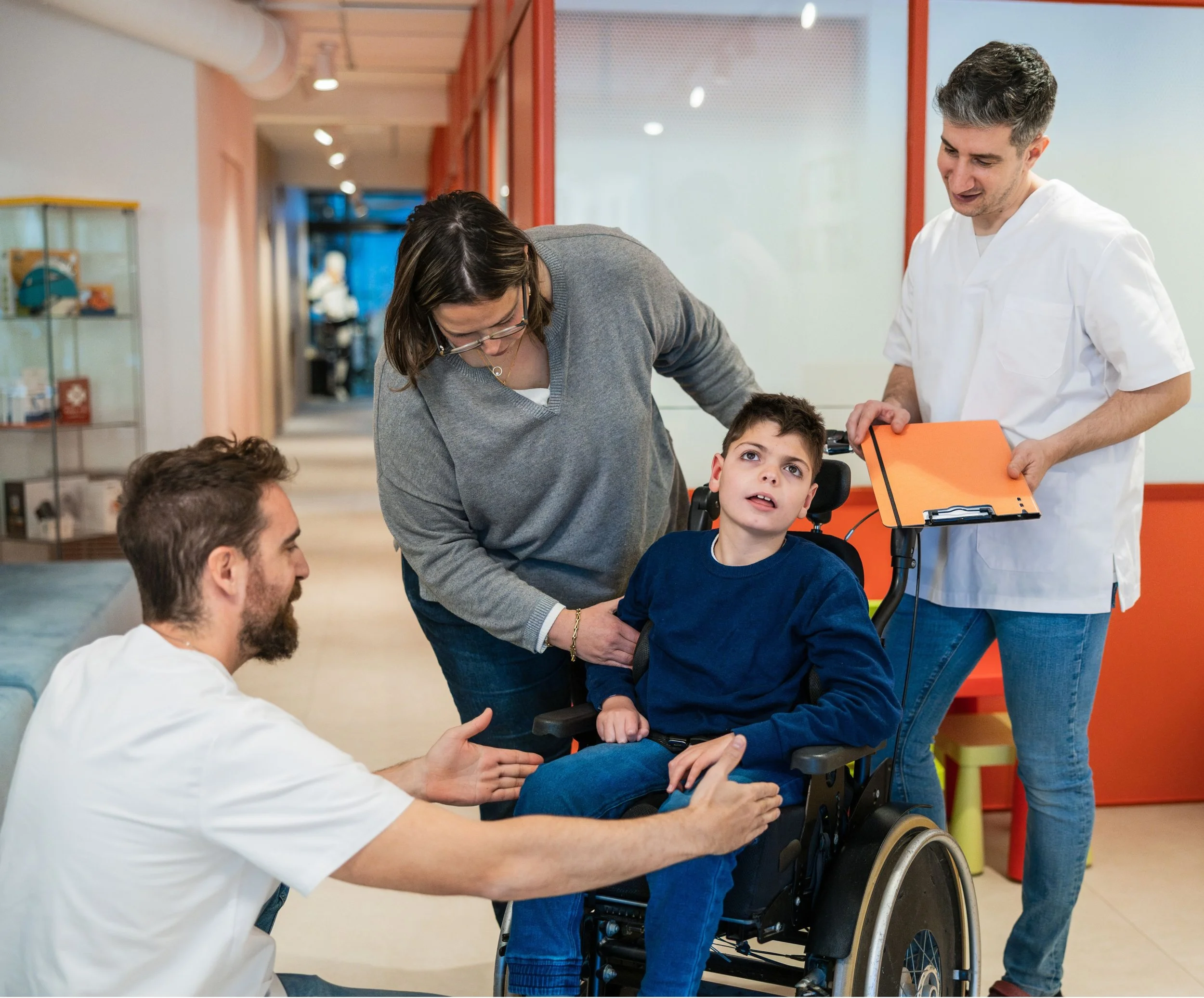 A healthcare professional kneels beside a young boy in a wheelchair and shows him something while another healthcare professional stands nearby holding a clipboard in a brightly lit hospital corridor.