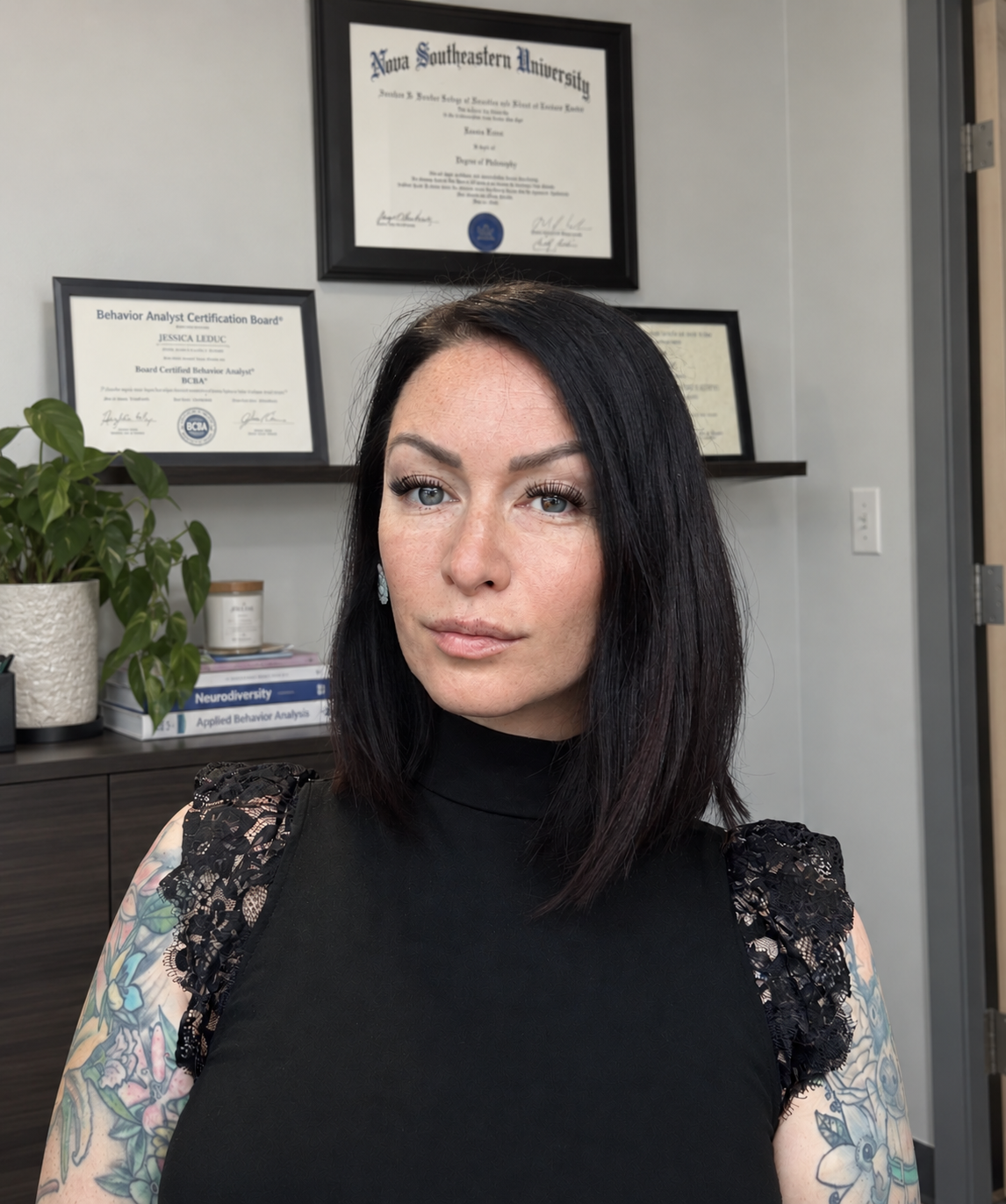 A woman with black hair and tattoos on her arms, wearing a black lace shoulder top, standing in front of framed certificates on a wall.