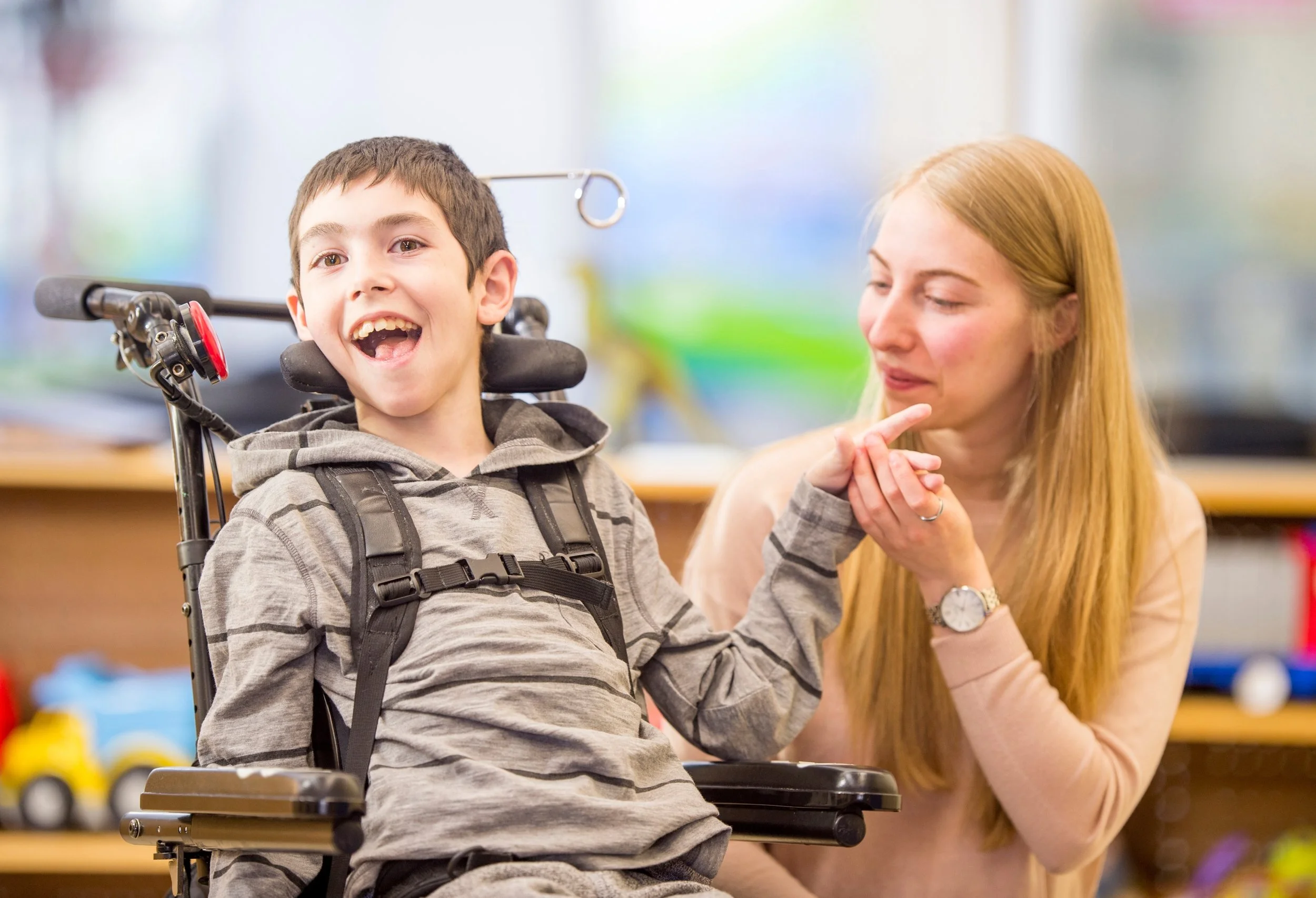 A young boy in a wheelchair smiling and talking to a woman with long red hair, in a colorful room.